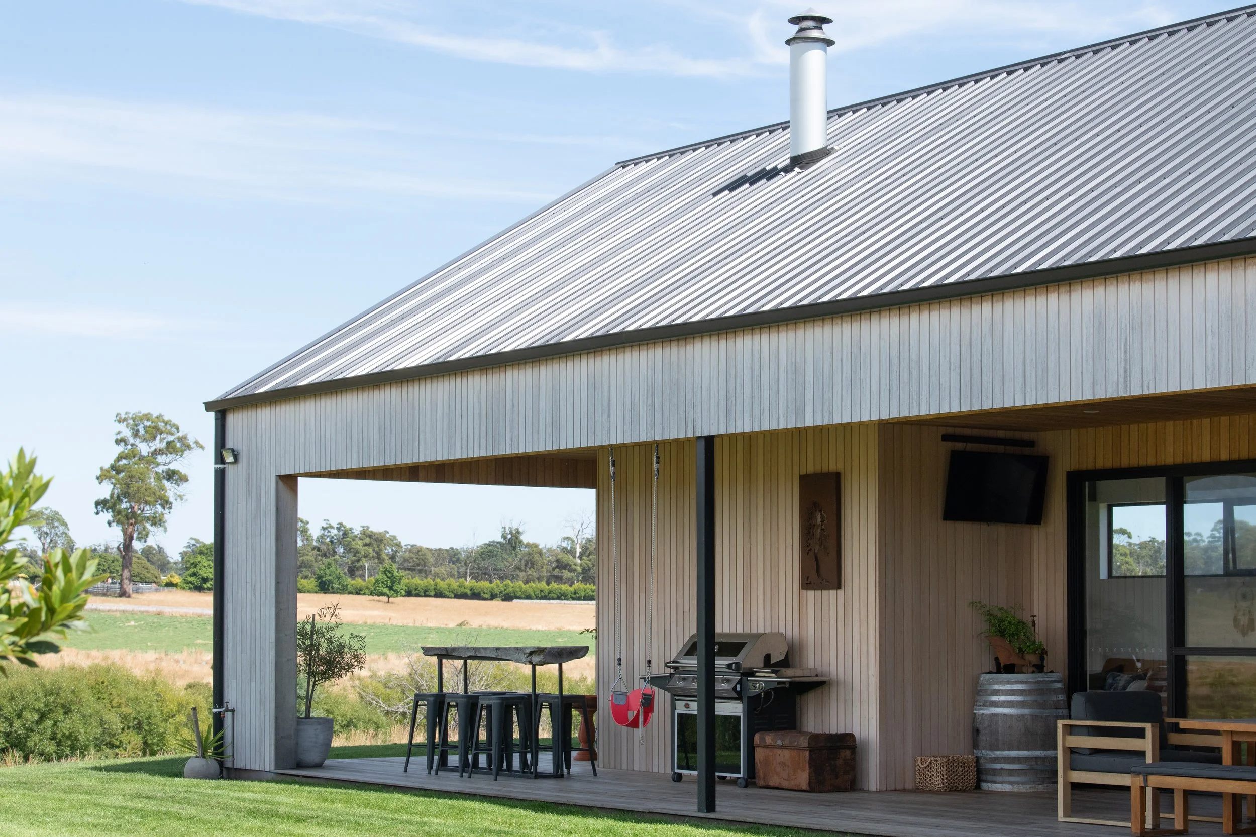 A modern barn house with a metal roof, outdoor grill, and patio, surrounded by trees and open fields under a blue sky.