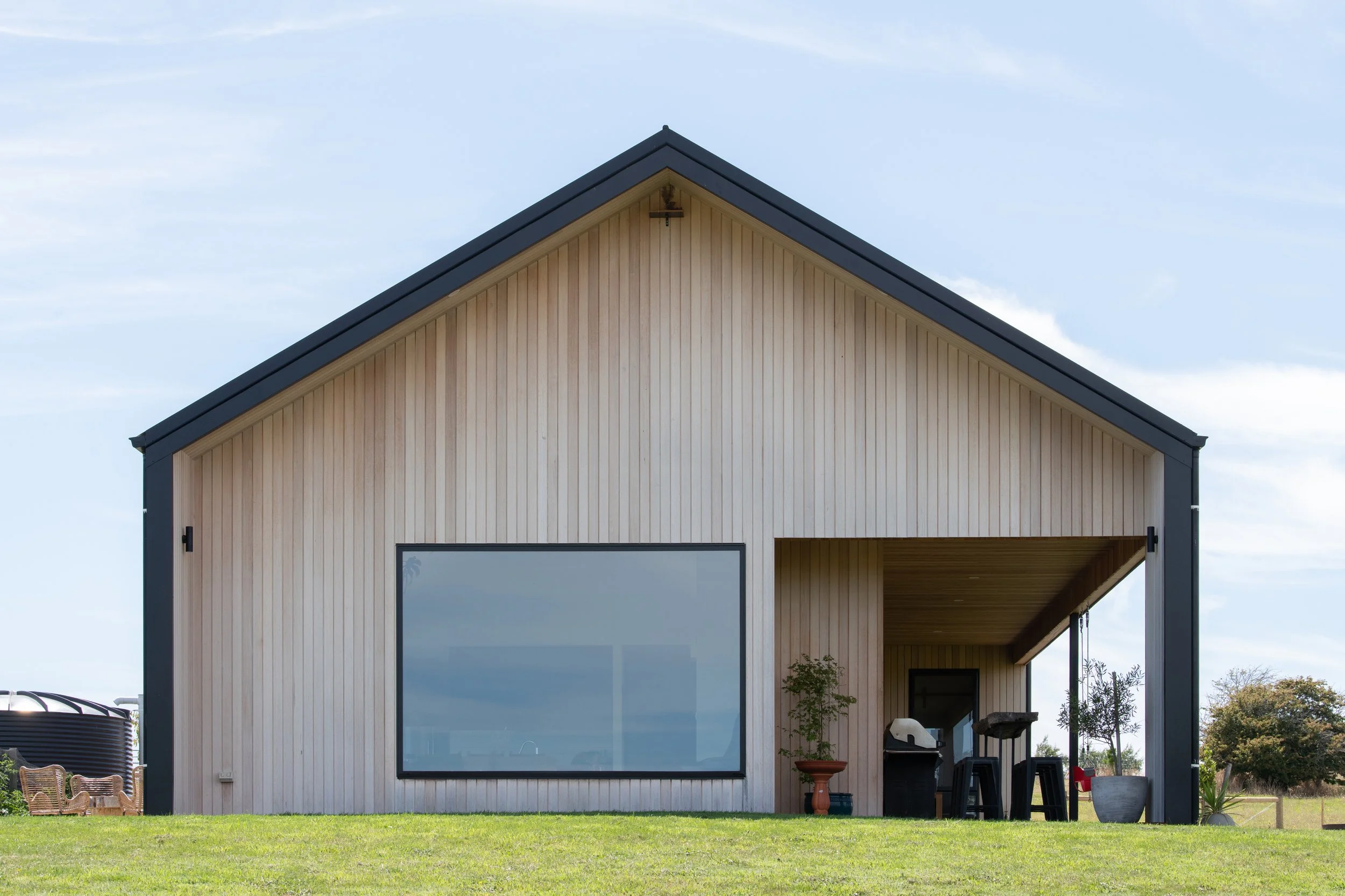 Modern house with a gabled roof, large glass window, and a covered patio in a grassy field.