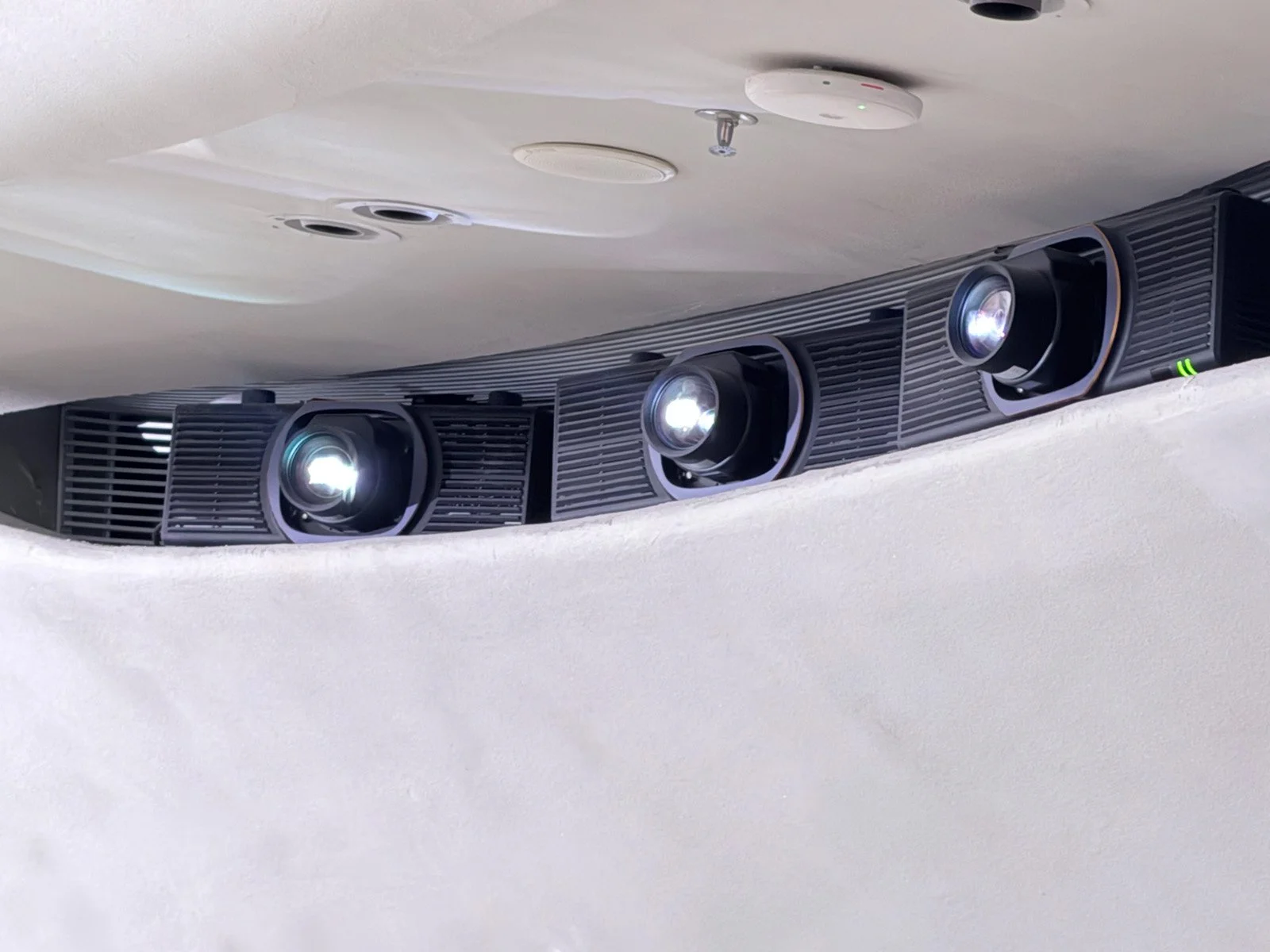 Three black modern projectors mounted on a white ceiling, with one turned on and emitting light.