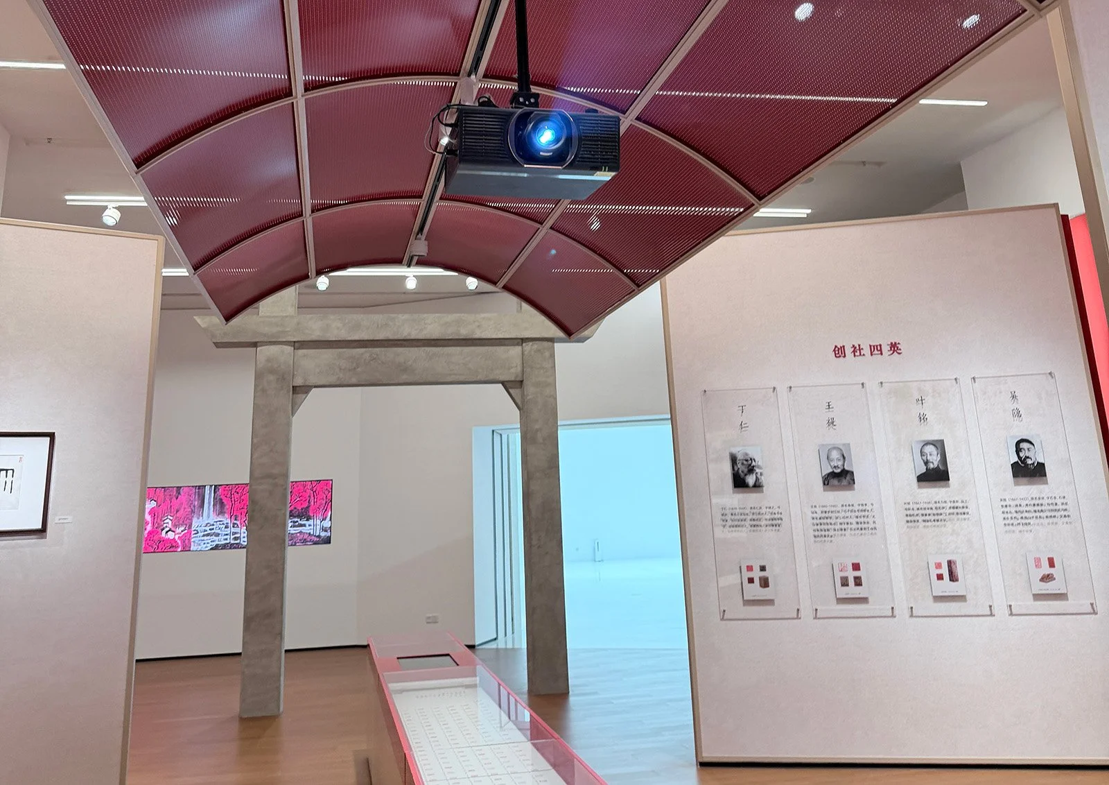 Interior of an art gallery featuring a red curved ceiling structure above a stone archway. There are informational posters on dividing panels, one displaying black and white portraits with Chinese text. In the background, there is a digital screen with pink and purple artwork and a white room visible through a doorway.