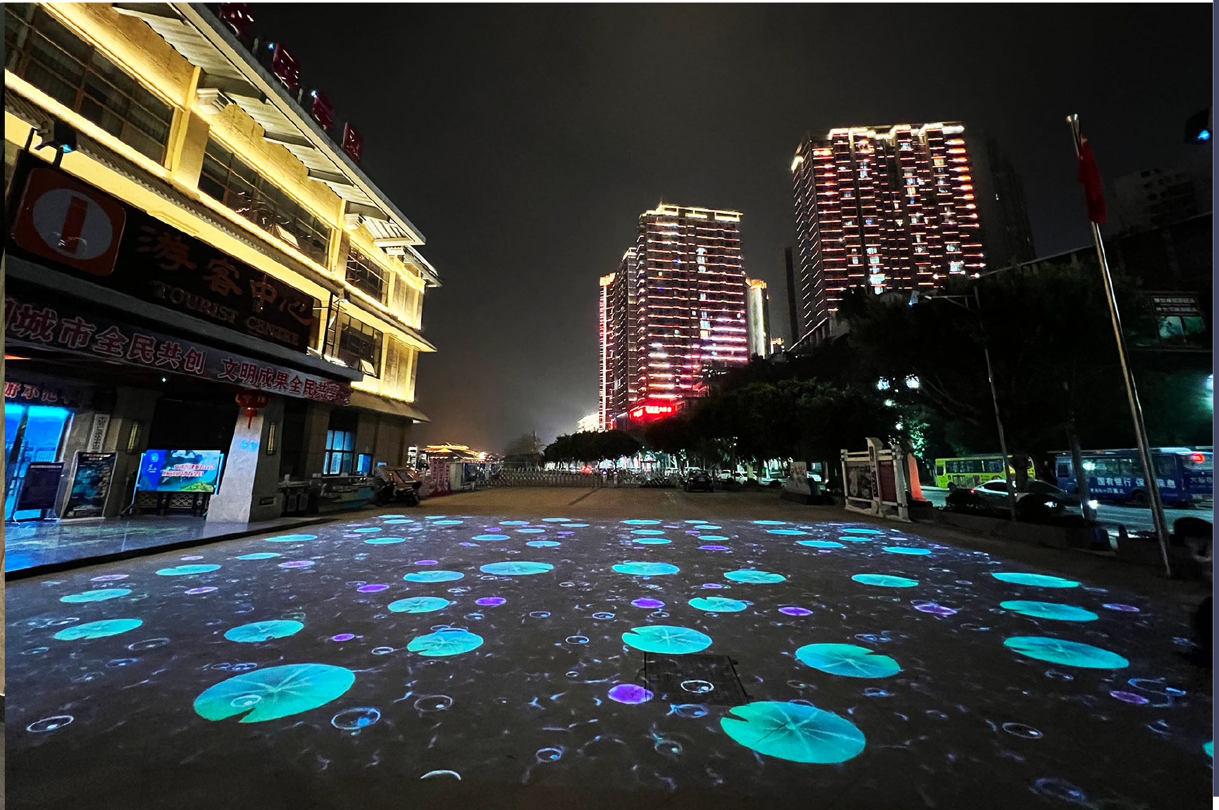 Night view of a city street with tall buildings illuminated with colorful lights, and a decorative light projection of glowing circles and patterns on the street pavement.