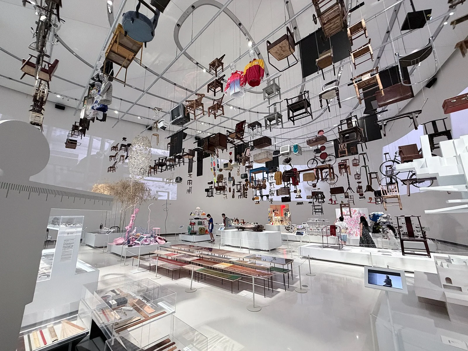An art installation featuring chairs and other objects hanging from the ceiling in a gallery space. Various chairs are suspended at different heights, creating a floating effect, with some clothing items also hanging among the chairs.