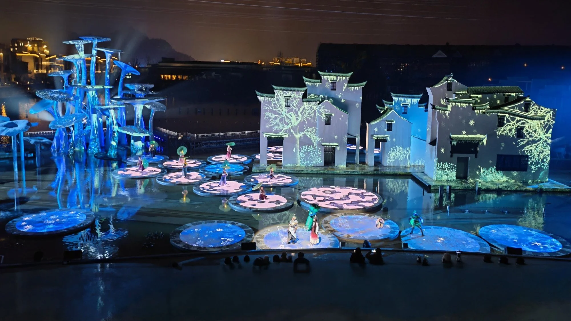 Nighttime performance in Wuyuan Cultural Tourism Town featuring illuminated traditional architecture, blue mushroom sculptures, and umbrella dancers.