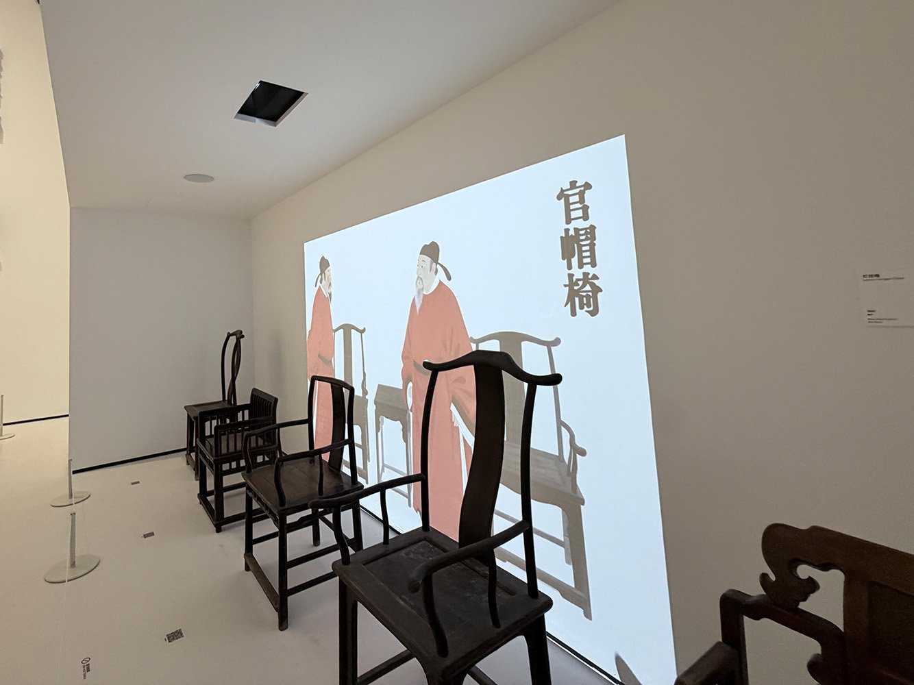 Exhibition room with traditional Chinese chairs and a digital projection of Chinese artwork featuring two historical figures and Chinese characters.
