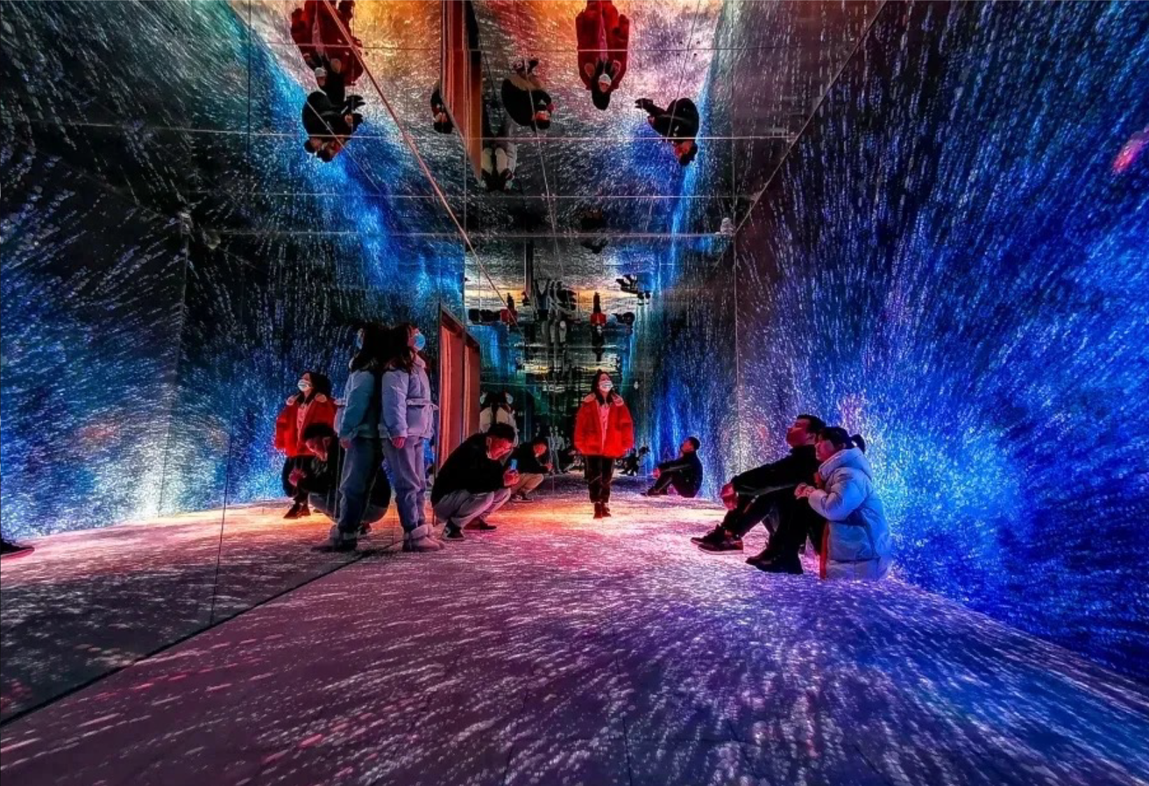 People inside a mirrored tunnel with colorful, starry light projections on the walls, ceiling, and floor, creating a vibrant, cosmic atmosphere.