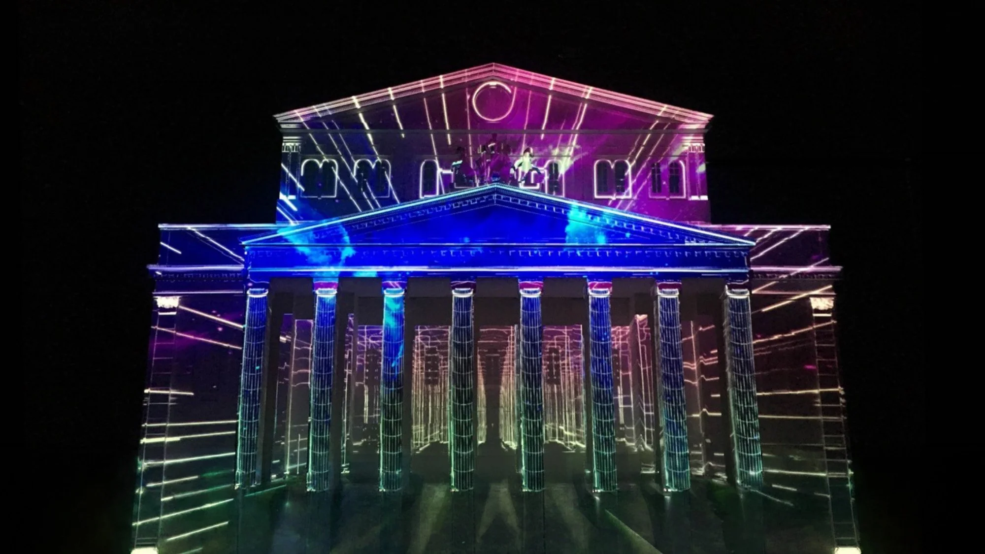 "Gate of Light" mapping show: Ancient Greek temple with columns illuminated in neon pink, blue, and green against night sky.