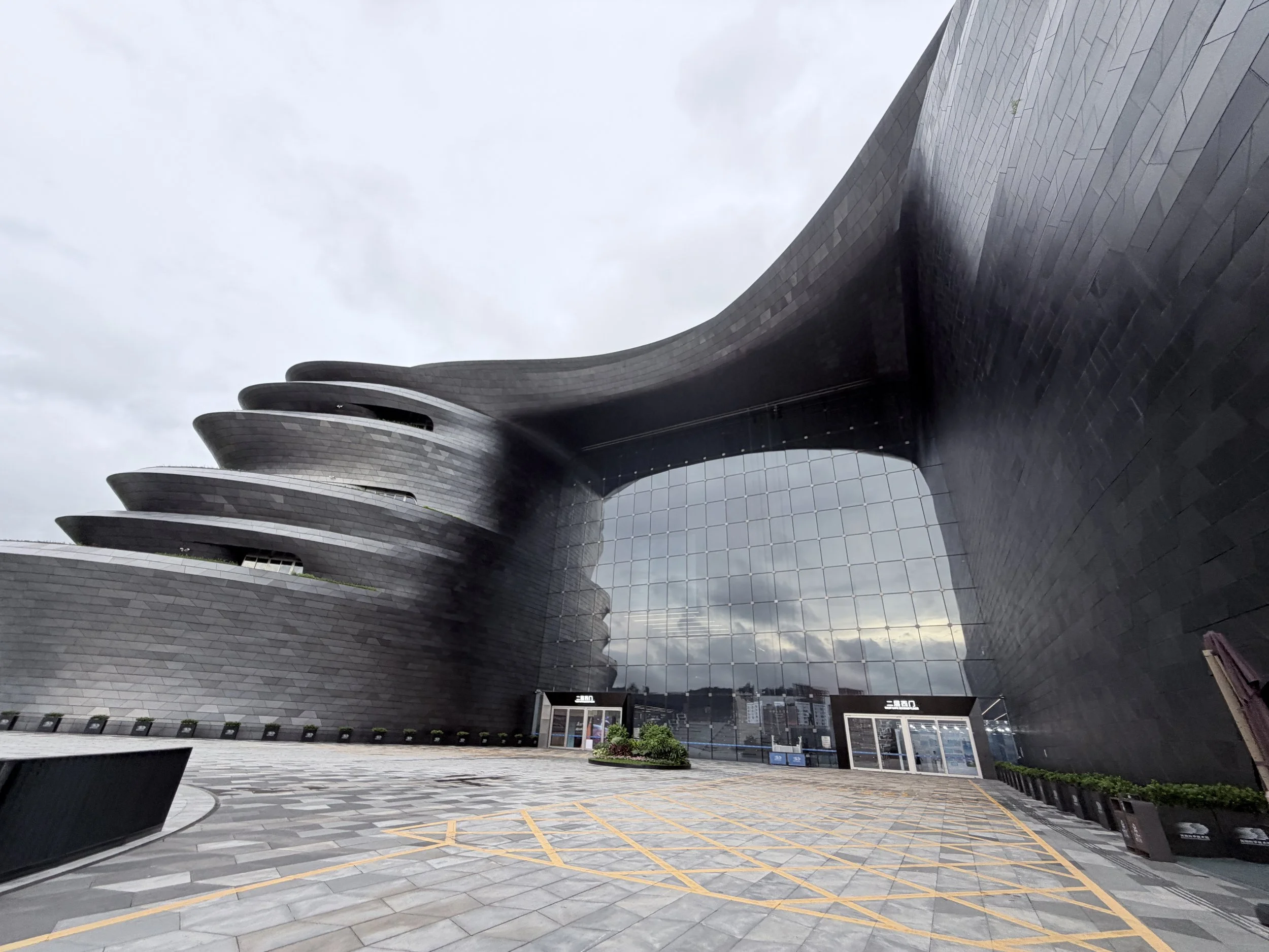 Modern high-rise building with a unique curved architectural design and reflective glass windows, with an open urban plaza in the foreground, featuring parking spaces and planters, against a cloudy sky.
