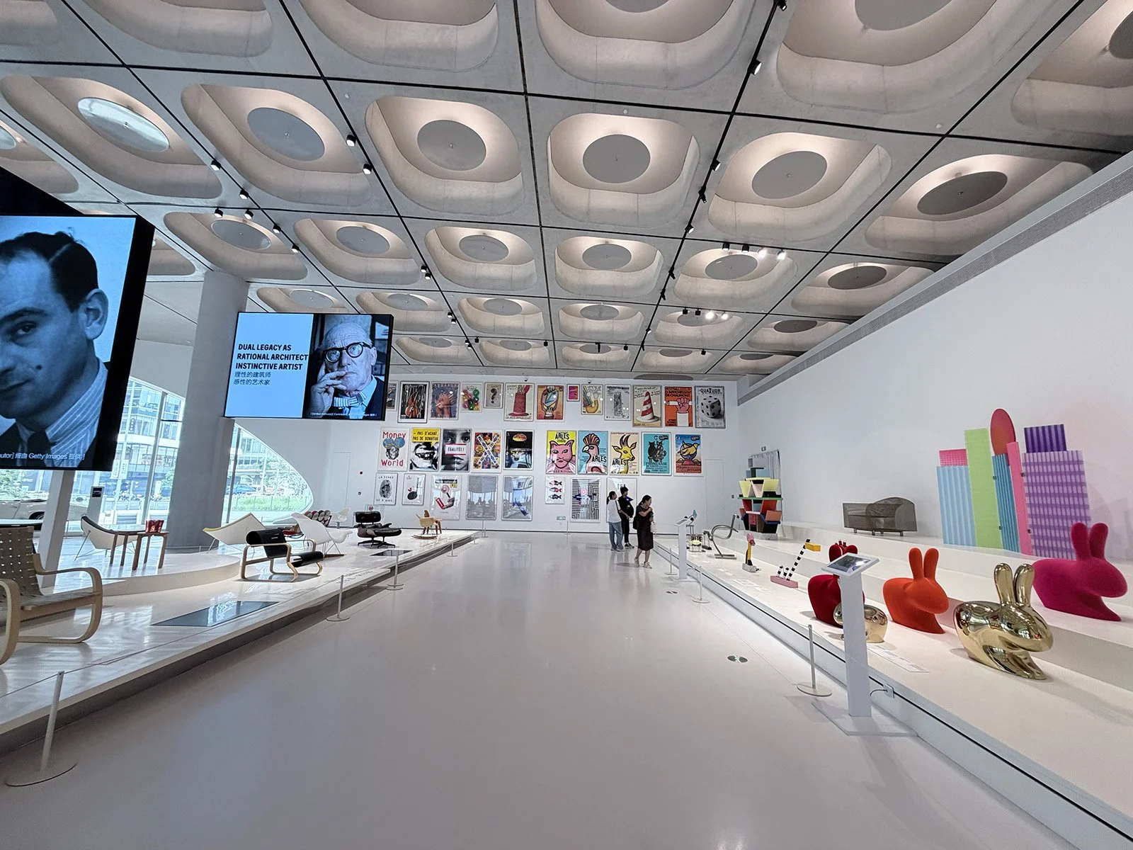 A modern art gallery with various colorful artworks, sculptures, and furniture displays, including chairs and rabbit-shaped objects, with visitors observing the exhibits.