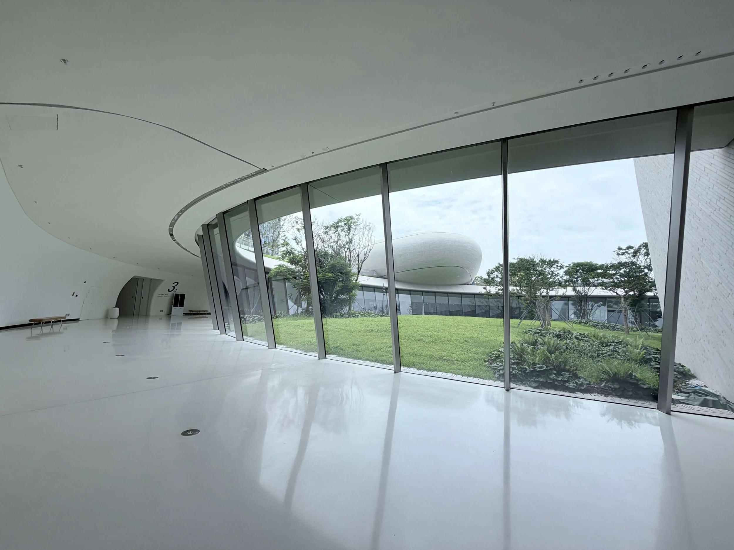 Shenzhen Cultural Square: Interior view through large glass windows facing an outdoor grassy lawn with trees.