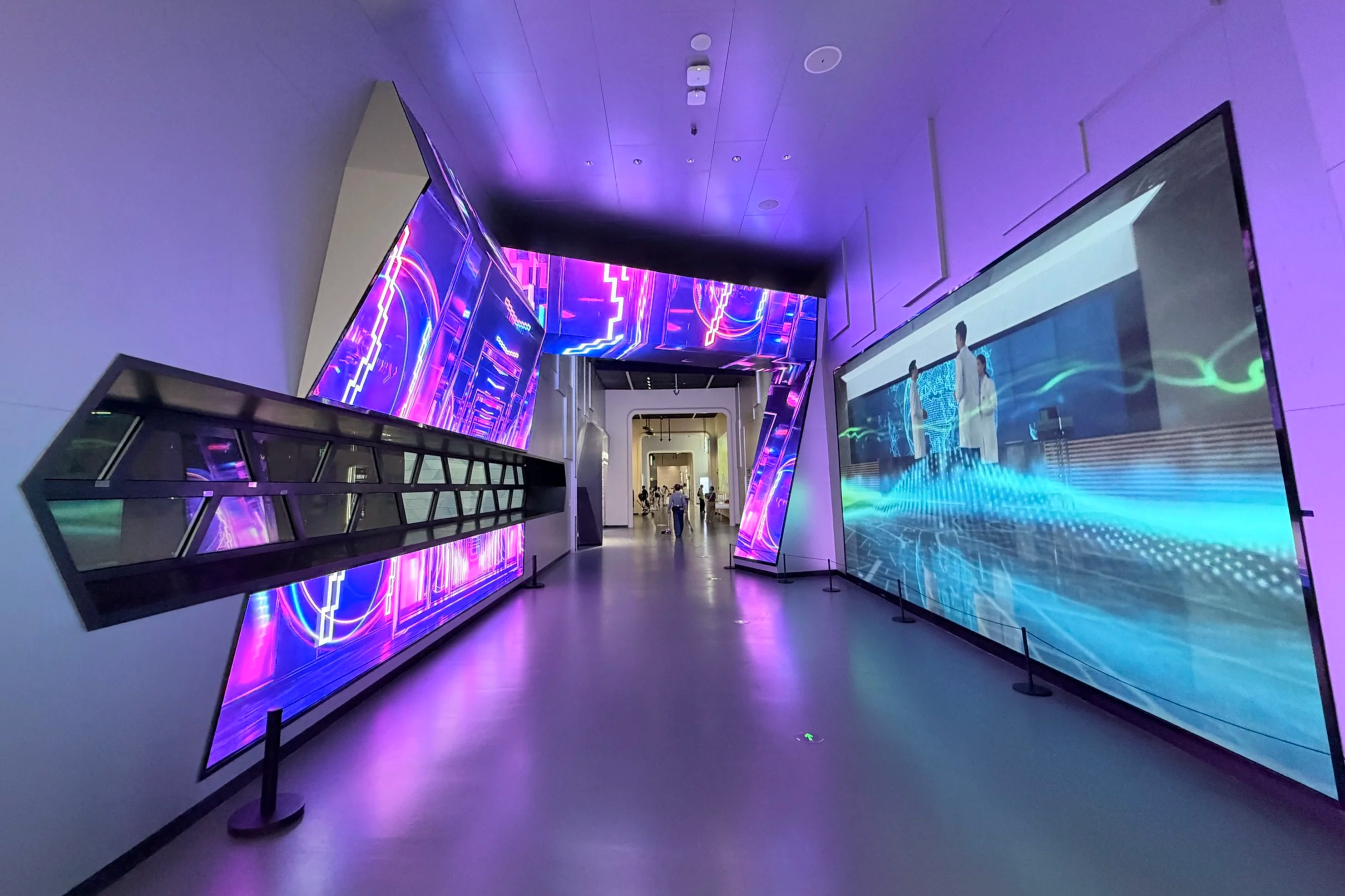 An interior view of a modern exhibition hall with large digital screens displaying colorful abstract graphics and visuals, illuminated by purple and blue lighting.