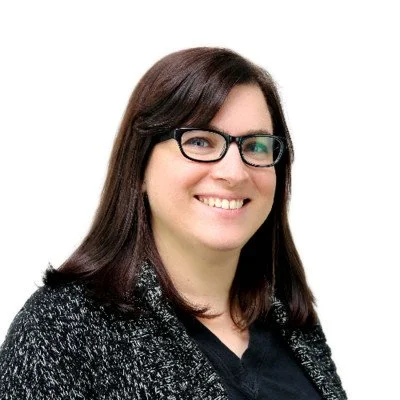 A woman with shoulder-length dark brown hair, wearing glasses and a black outfit with a black textured sweater, smiling against a plain white background.