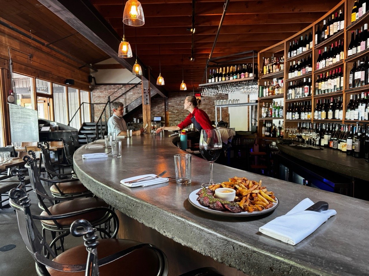 View of a bar with a curved concrete countertop, wine bottles on shelves, a glass of red wine and a plate of steak and fries on the counter, and a bartender serving a customer.