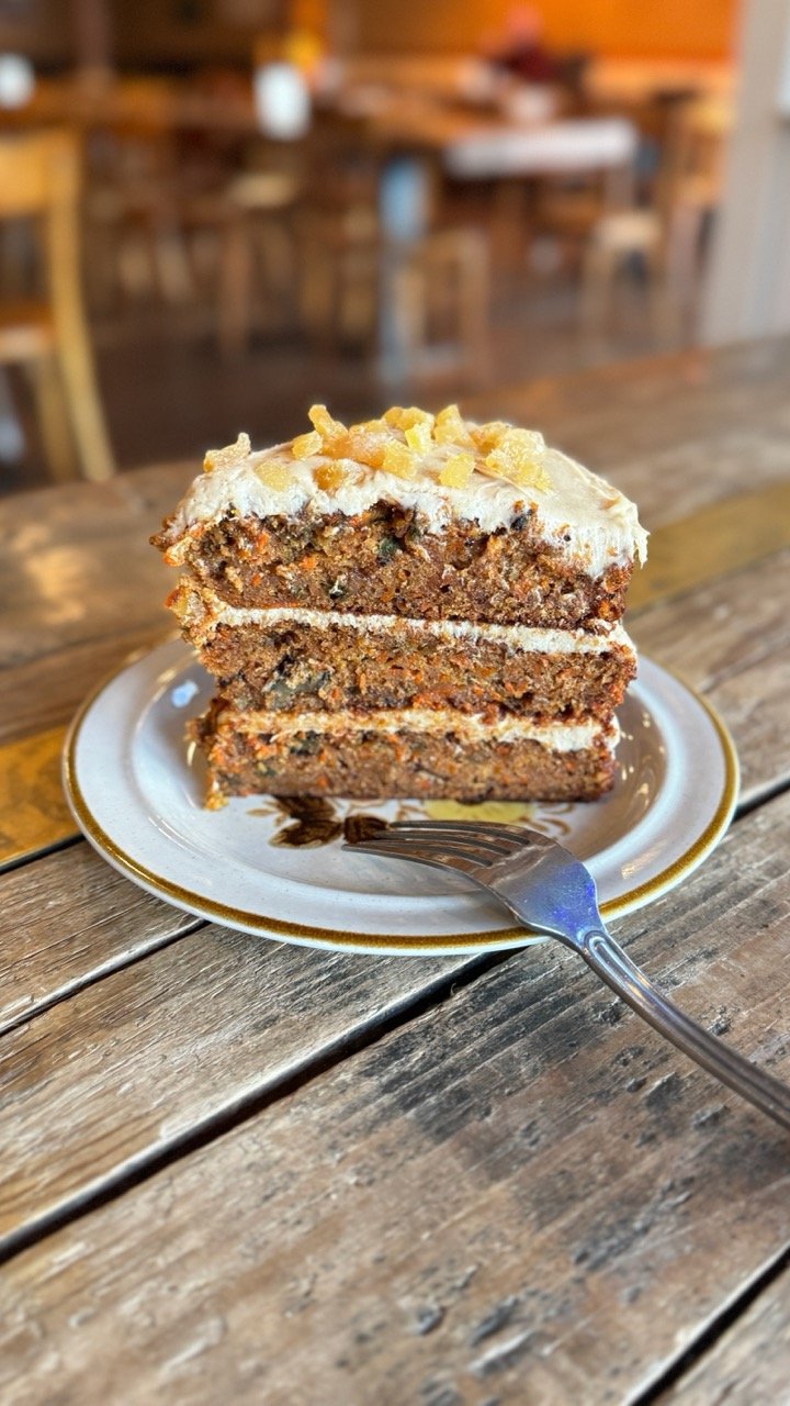 A slice of carrot cake with cream cheese frosting and candied ginger on top on a decorative plate, with a fork resting on the plate, on a rustic wooden table in a cozy restaurant or café setting.