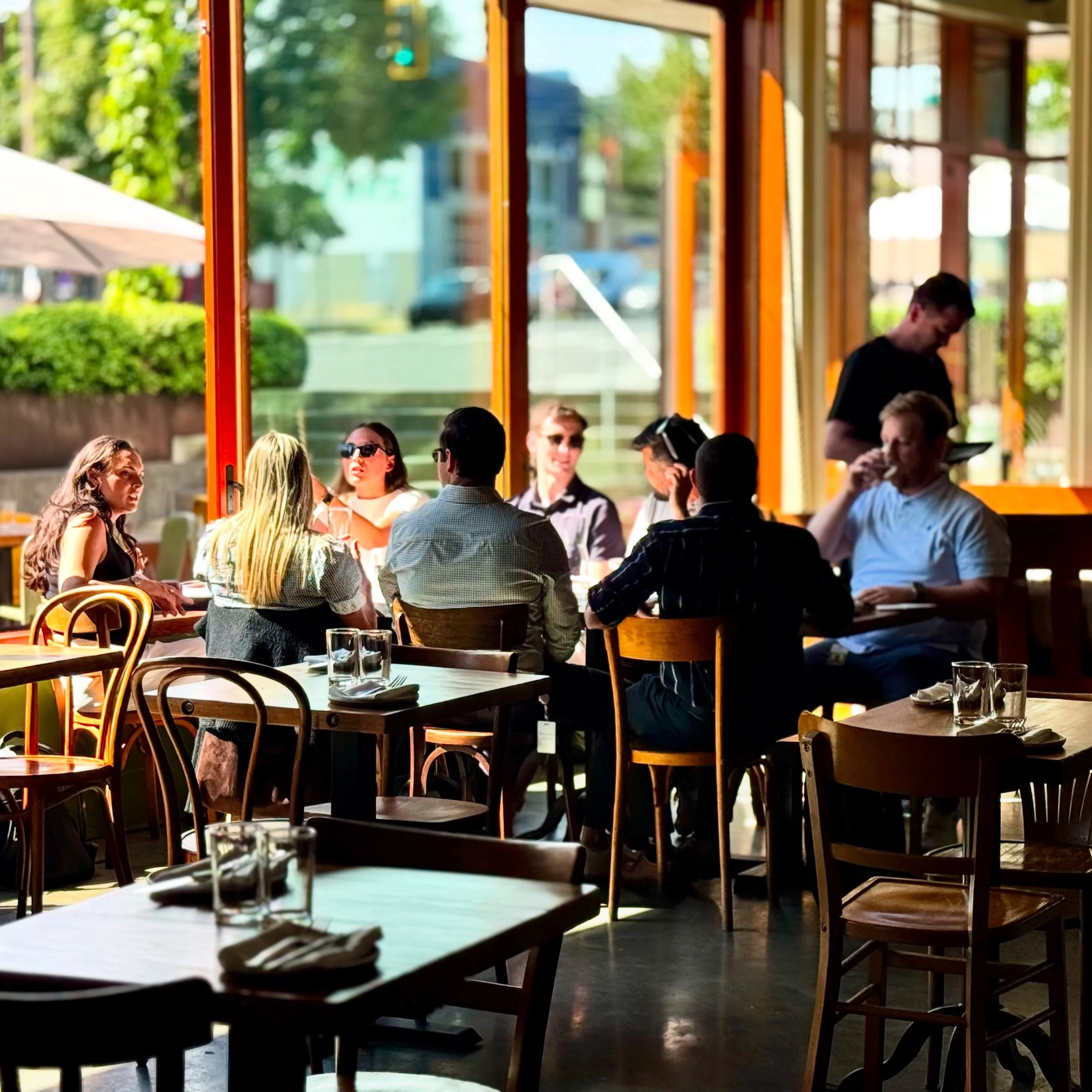 People dining inside a restaurant with large windows, sunlight streaming in, and green outdoor scenery visible outside.