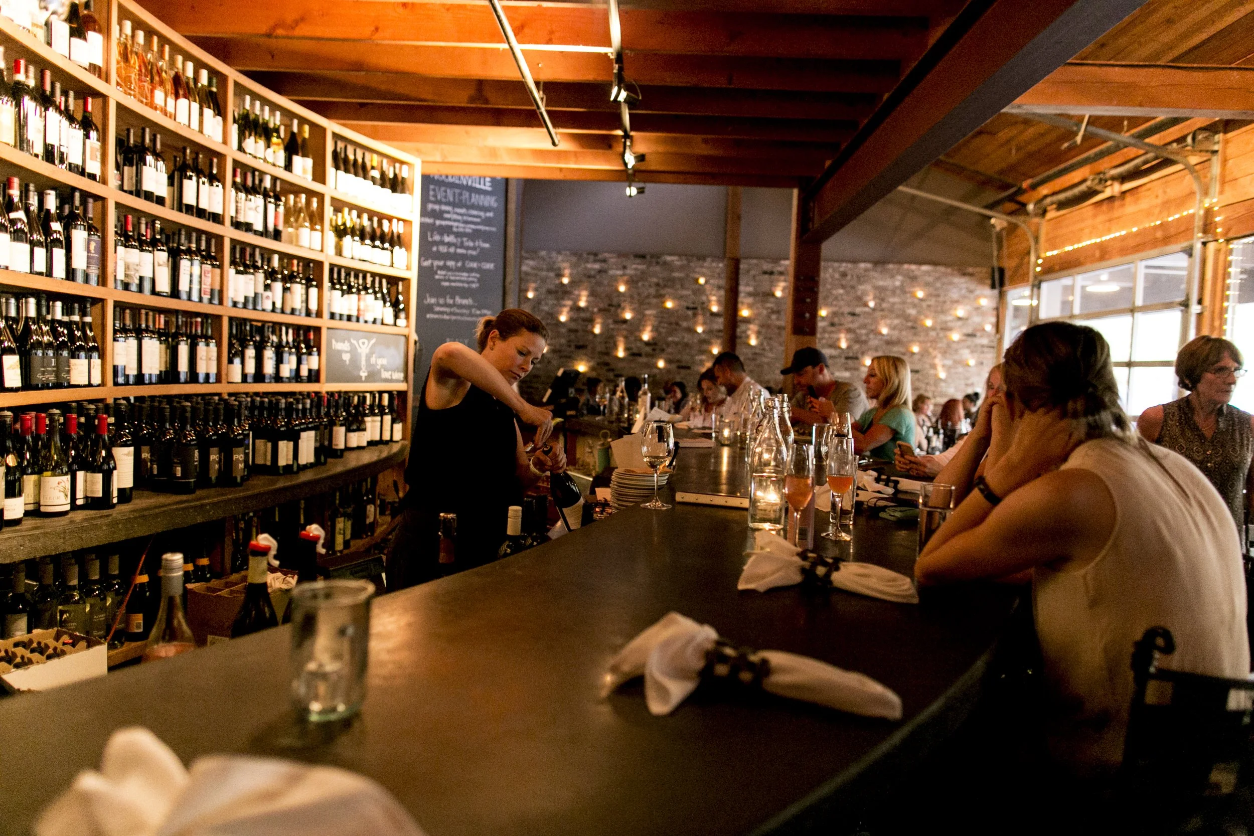 Inside a cozy restaurant or bar with brick walls and wooden beams, a bartender preparing drinks behind the counter, and patrons sitting at the bar and tables, enjoying their meals and drinks.