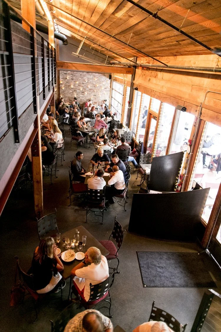 People dining in a cozy restaurant with wooden ceiling, stone wall, and large windows, enjoying meals and drinks.