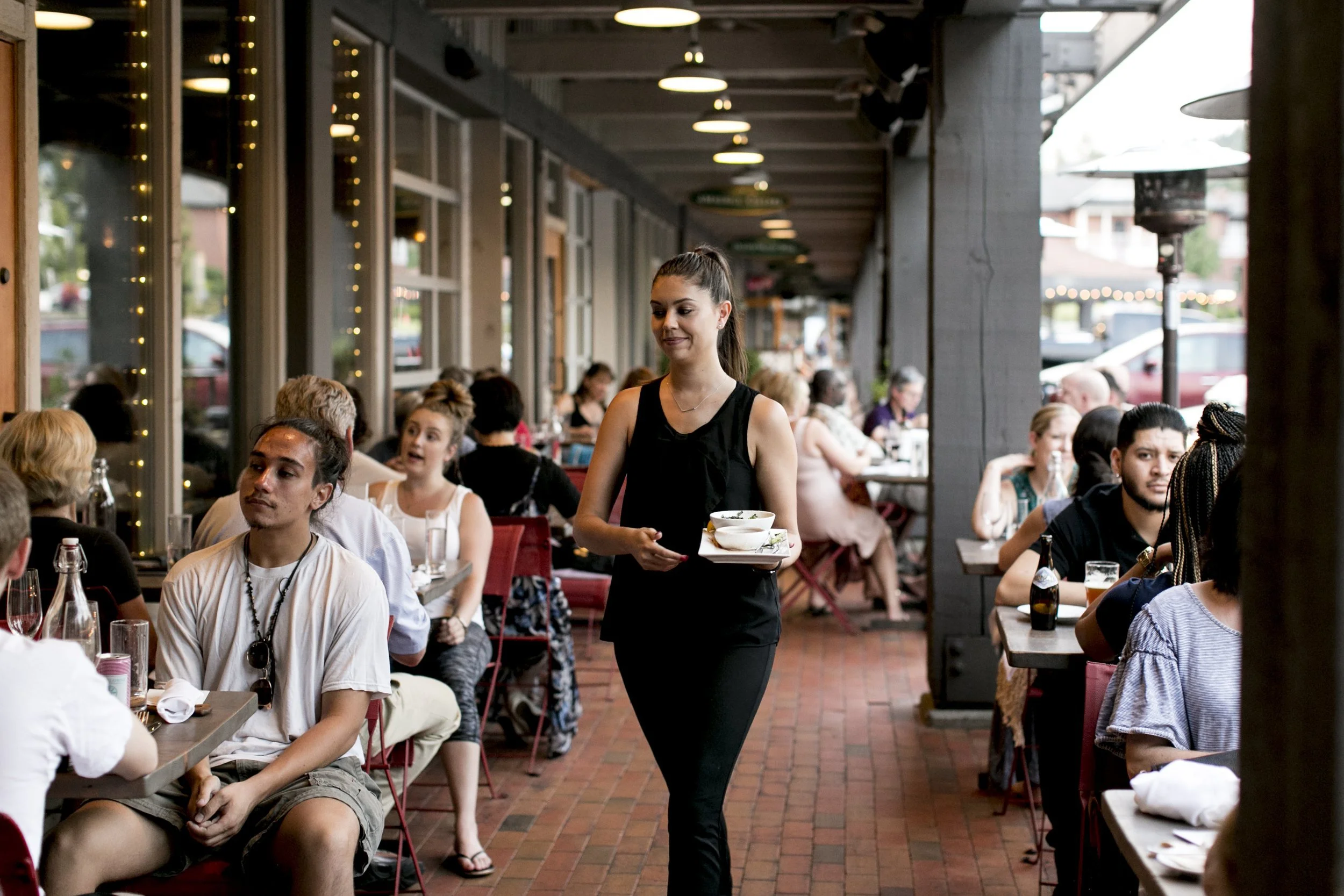 A young woman in a black dress is serving drinks to a group of people dining at an outdoor restaurant or patio during daytime.
