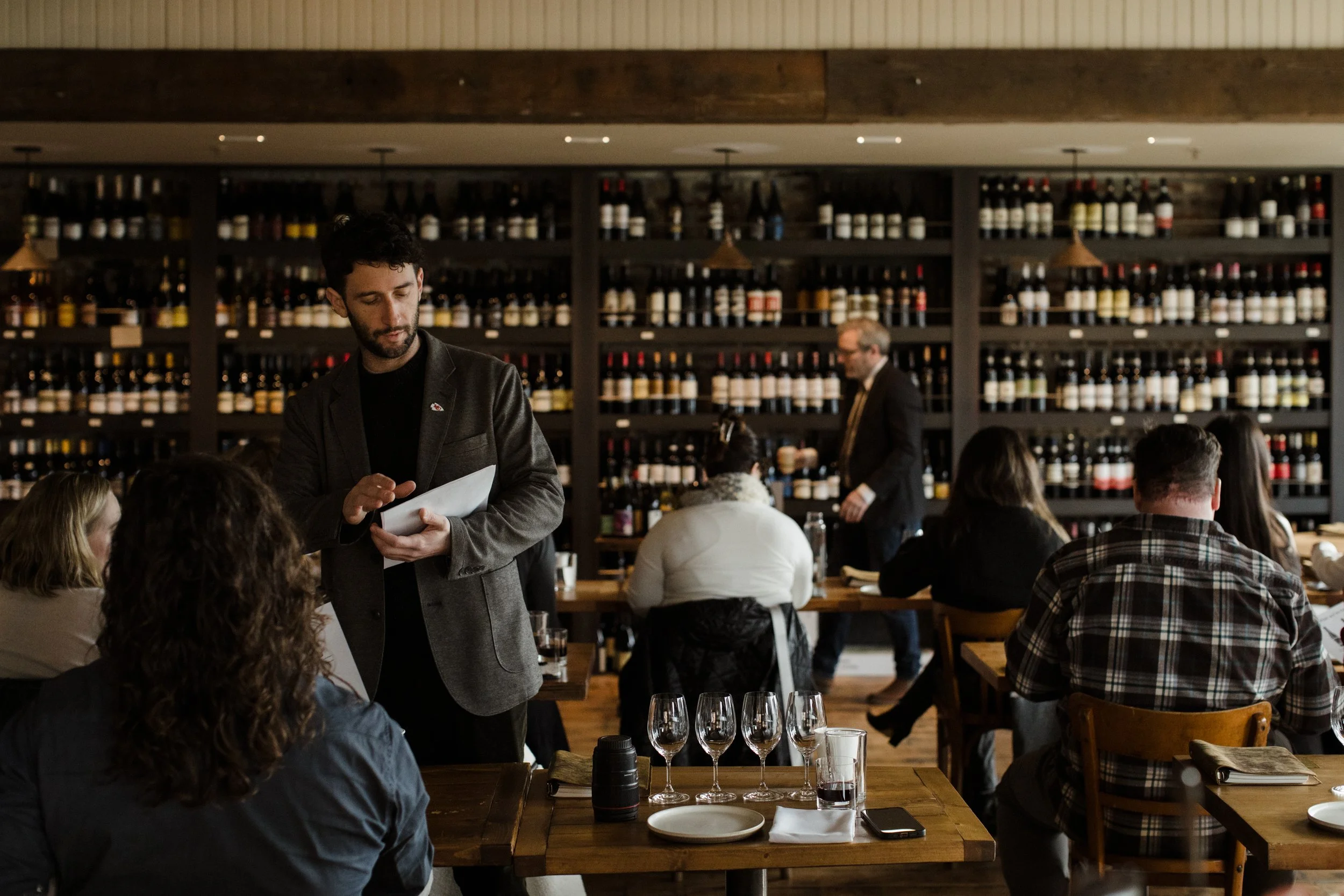 A waiter in a gray blazer is taking an order at a busy restaurant with wine bottles on wooden shelves in the background, and customers sitting at wooden tables.