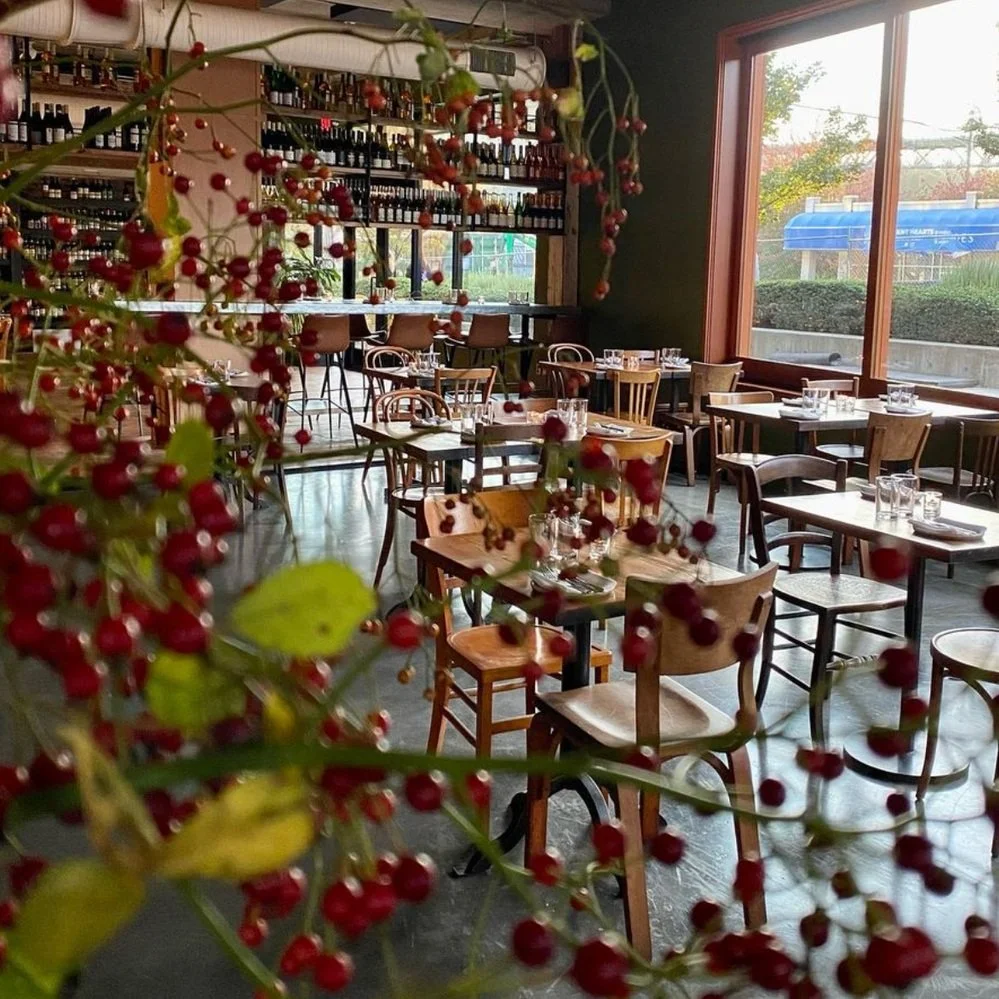 Inside a cozy restaurant with large windows, wooden tables and chairs, and a view of trees outside. A bunch of red berries and green leaves partially obstruct the view.