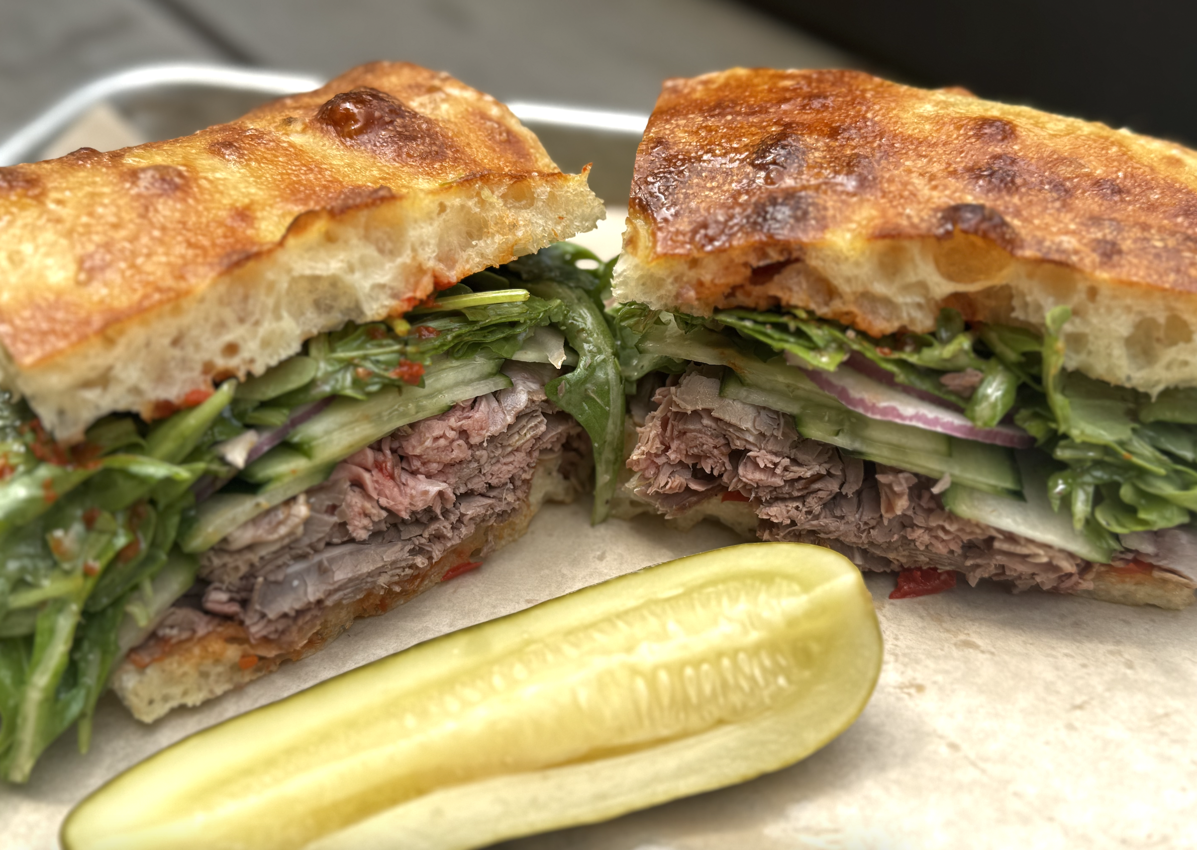 Close-up of a sandwich with toasted bread, shredded meat, lettuce, tomato, and pickles, with pickles on the side.