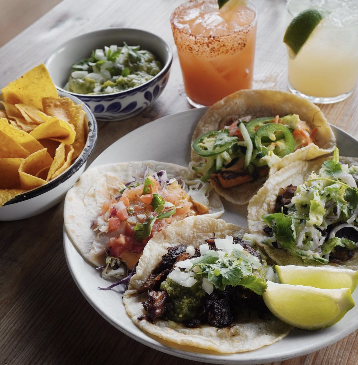 A plate of four tacos with various fillings, a bowl of tortilla chips, a bowl of guacamole, and two drinks, one with a lime slice and one with a lime wedge, all on a wooden table.