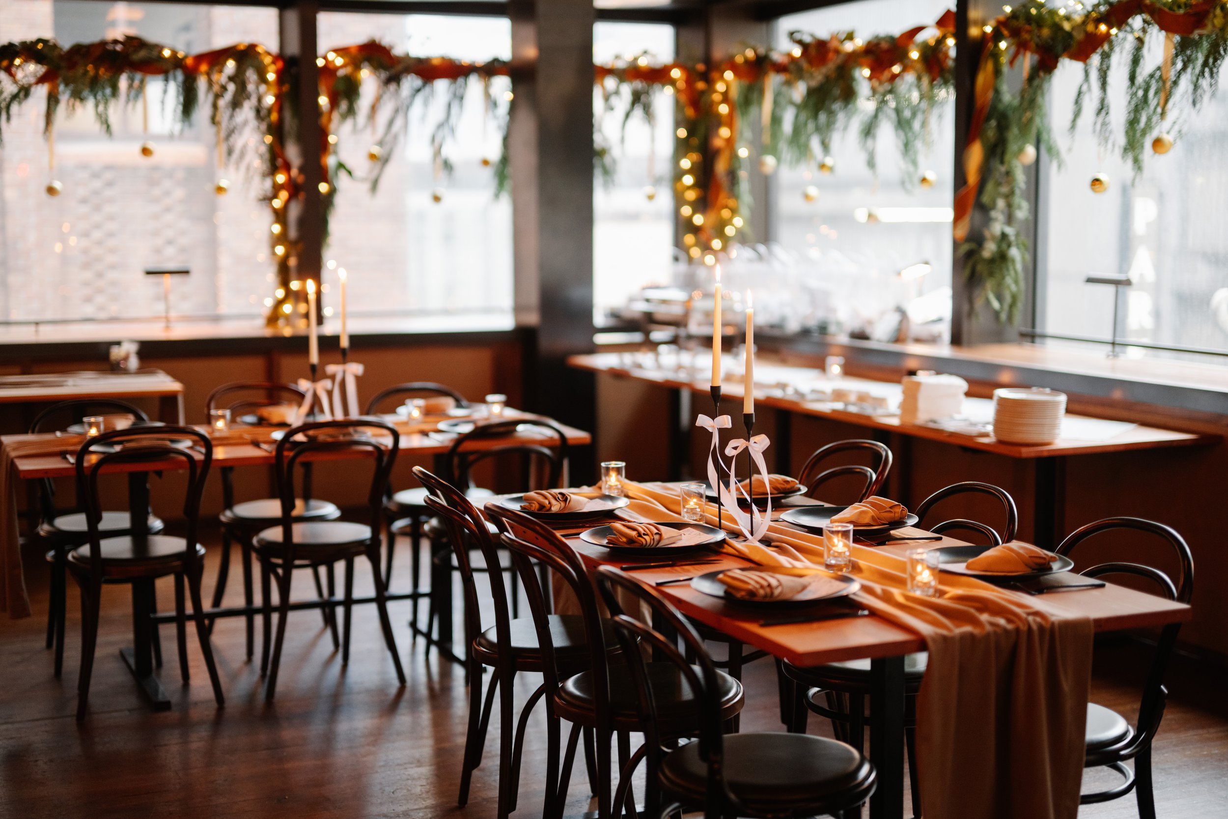 Decorated indoor dining area with tables set for a celebration, featuring tall candleholders with white ribbons, napkins, and glassware, and a backdrop of windows and hanging greenery with string lights and ornaments.