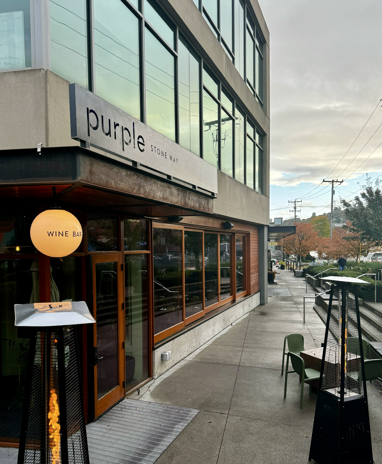 An urban sidewalk scene featuring a modern building with large glass windows, a sign reading 'Purple Stone Way,' and a restaurant with a warm, glowing sign that says 'Wine Bar.' Outdoor tables and chairs are visible on the patio, with heaters and som