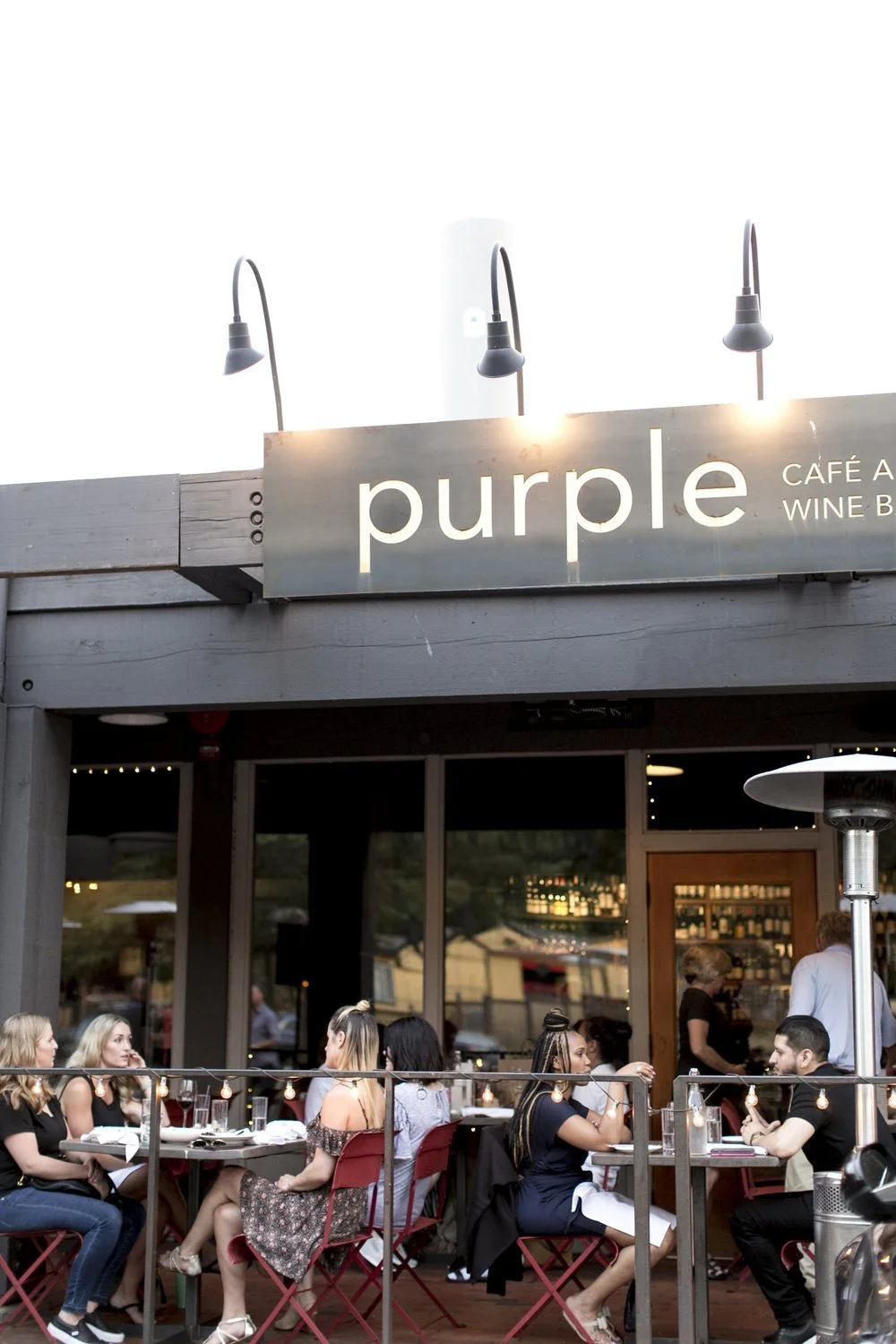 Outdoor dining scene at a restaurant called Purple, with people sitting at tables, engaging in conversations, outside of a cafe with warm outdoor lighting and a bar in the background.