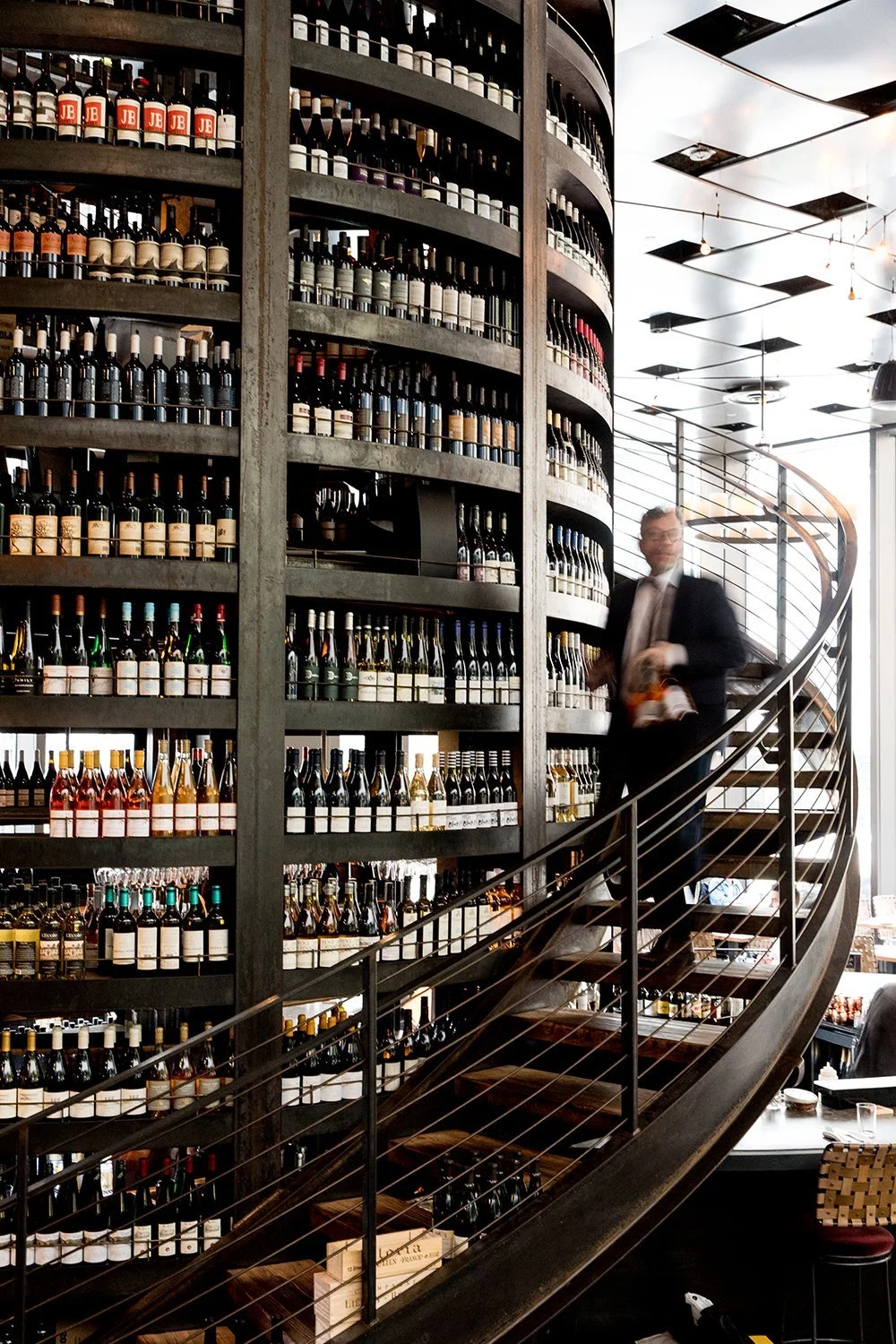 A spiral staircase next to a large wall of wine bottles on shelves, with a man in a suit walking down the stairs in a modern wine shop or restaurant interior.