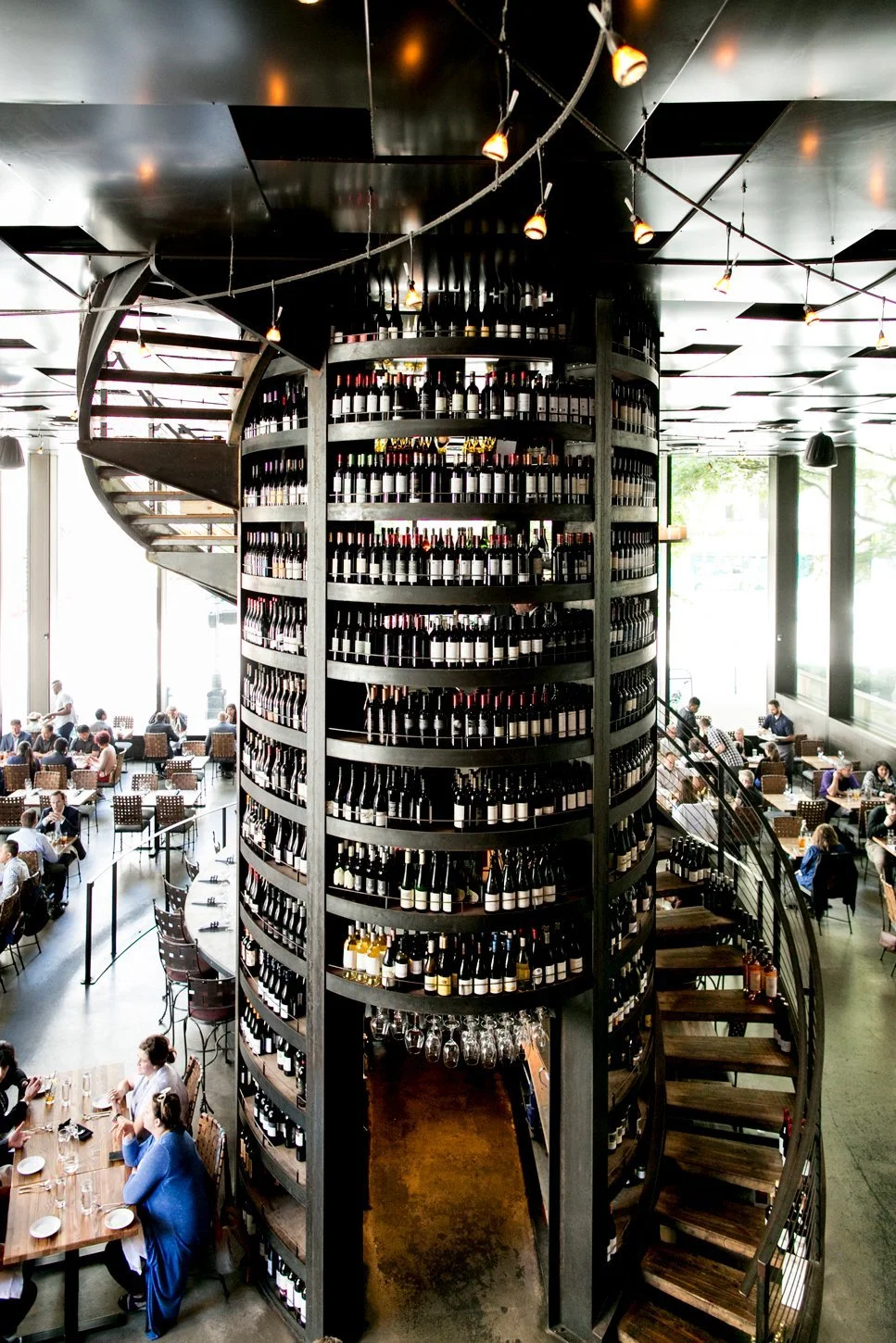 A large circular wine rack filled with bottles of wine, located in a modern restaurant with many diners seated at tables.
