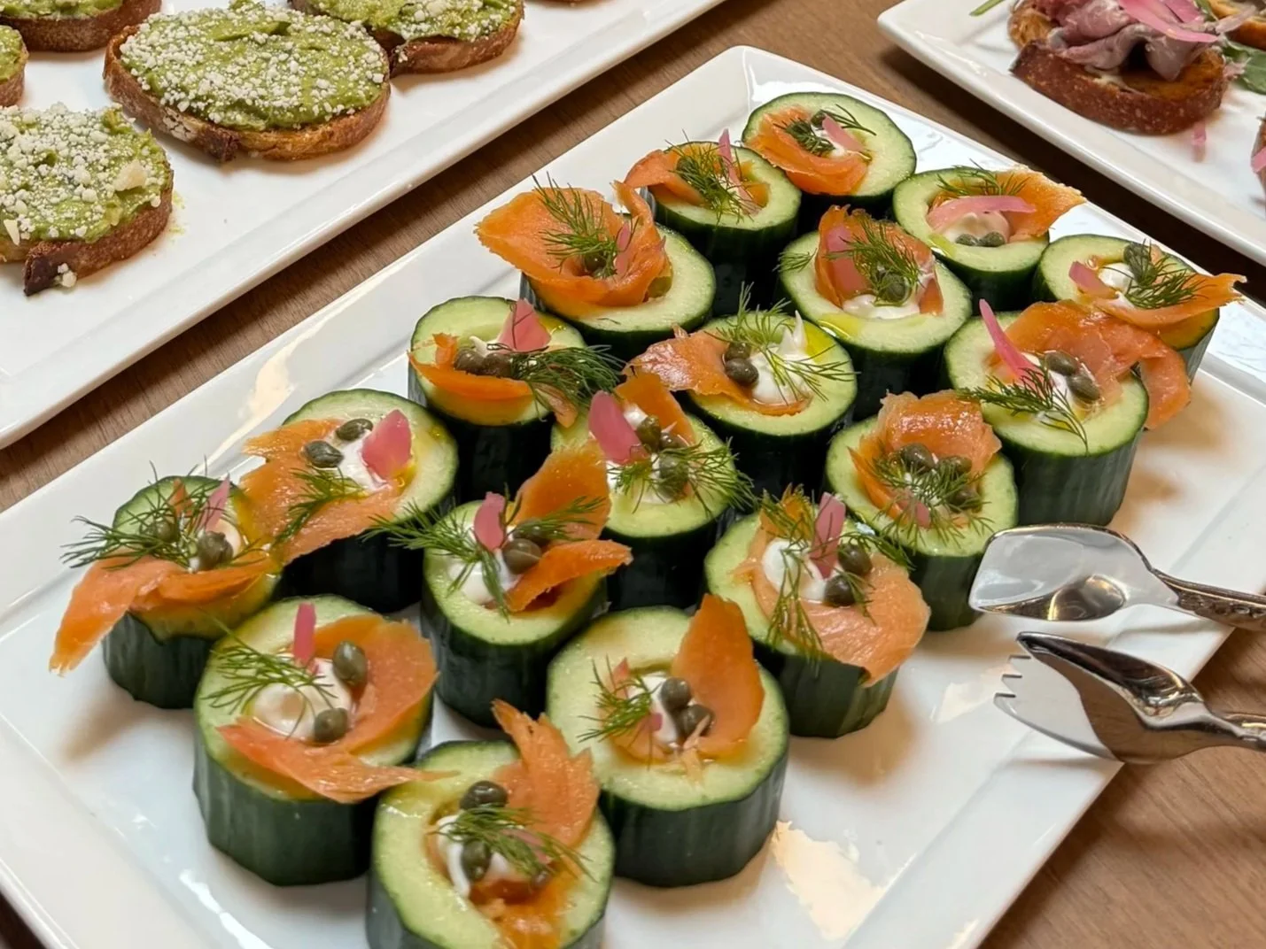 Cucumber slices topped with smoked salmon, cream cheese, caviar, dill, and pink pickled onions on a rectangular white platter.