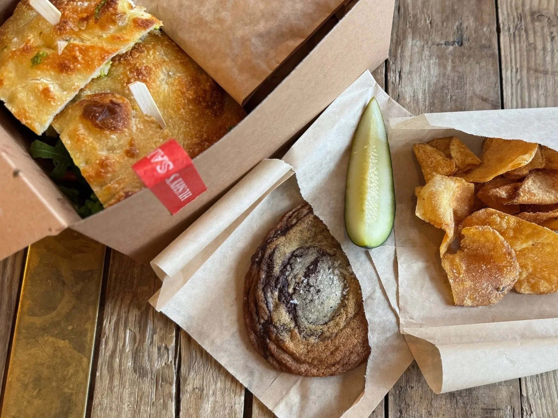 Assorted baked goods including a cinnamon roll, a slice of cucumber, fried chips, and a savory baked item in a brown paper tray on a wooden surface.