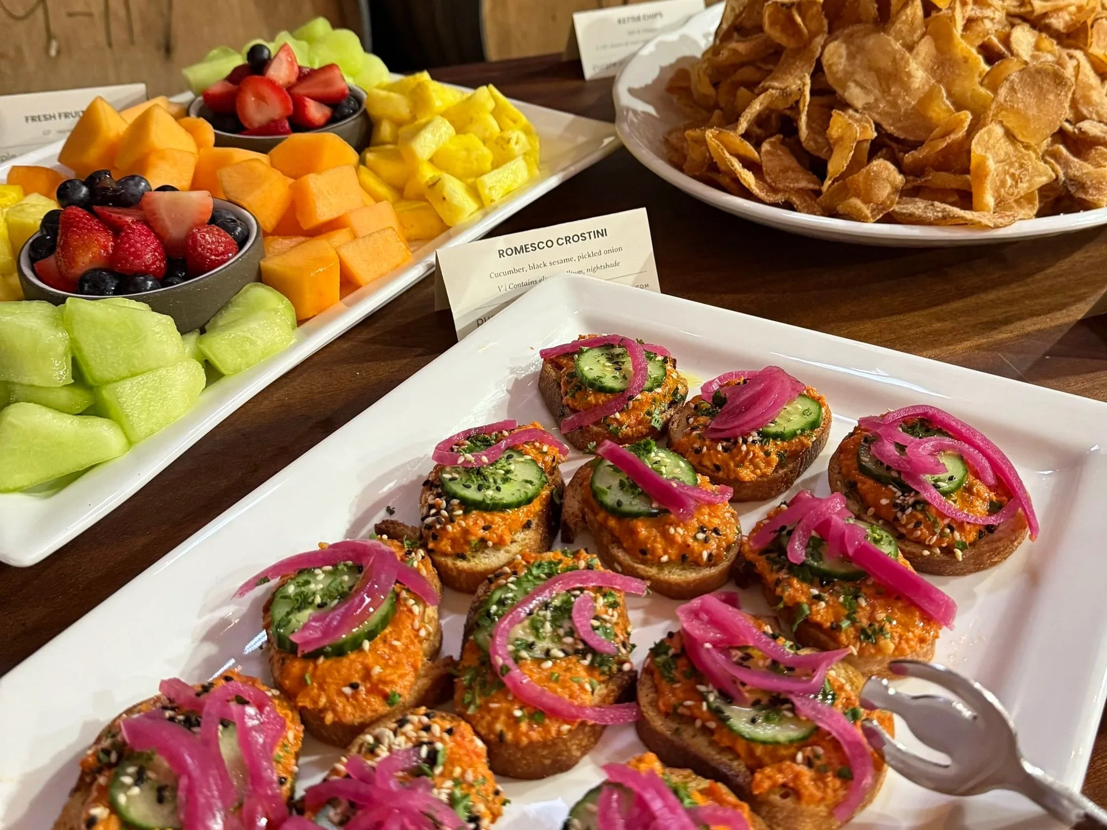 A platter of fresh fruit including strawberries, blueberries, cantaloupe, honeydew, pineapple, and watermelon, along with a plate of colorful appetizer toasts topped with cucumber, pickled onions, and herbs, and a plate of crispy fried food in the background.