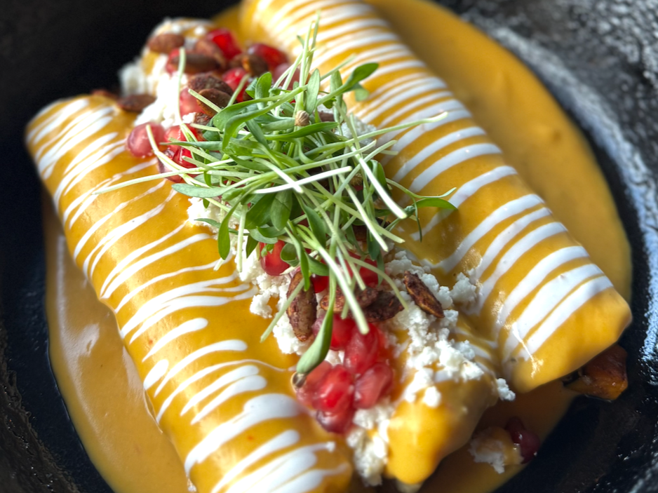 Two yellow Mexican tacos topped with white sauce, garnished with greens, pomegranate seeds, cheese, and grilled meat, served on a black plate.