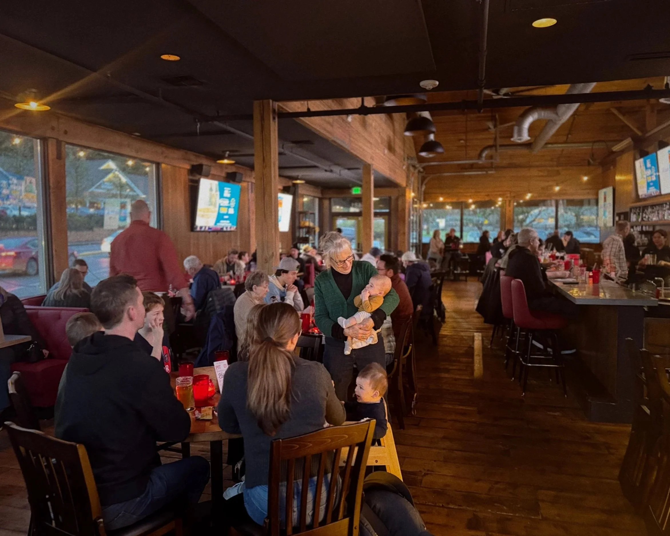 A busy restaurant with many diners, mostly seated at tables, some standing, and large windows showing an outdoor scene. The interior has wooden walls and ceiling, and dim lighting.