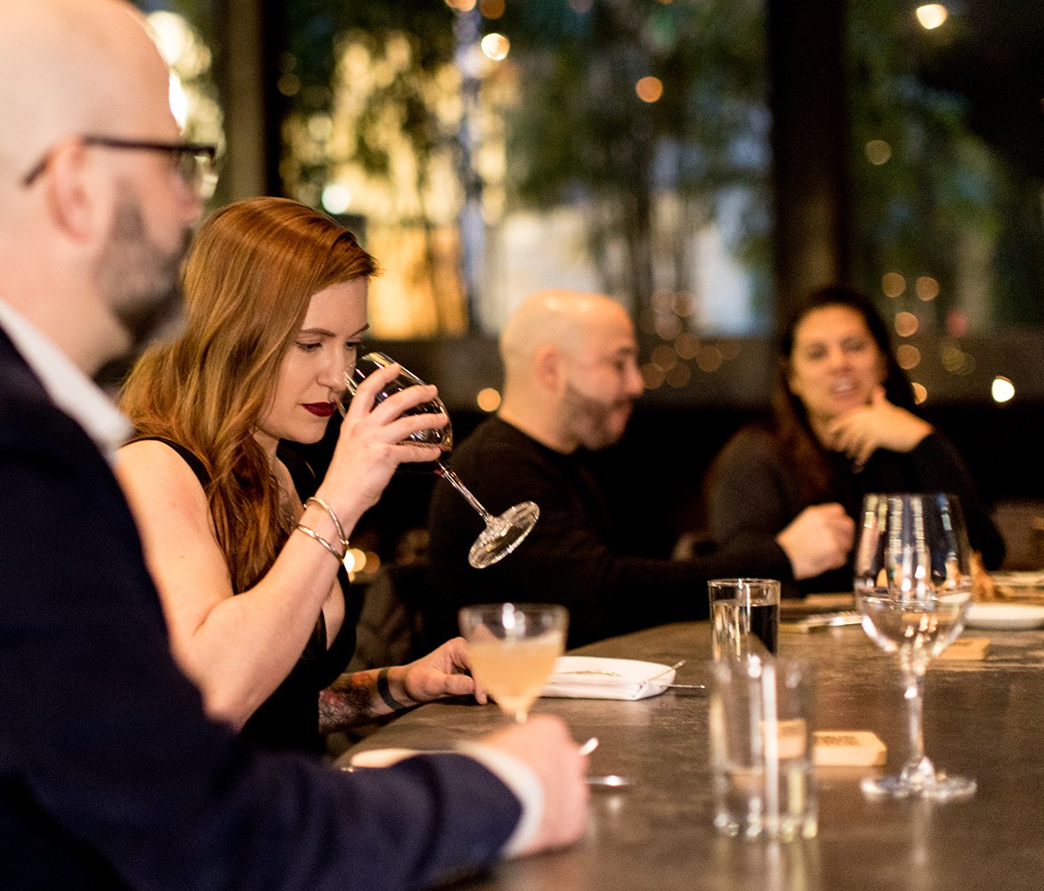 People sitting at a restaurant table, some with drinks, with a blurred background of lights and trees.