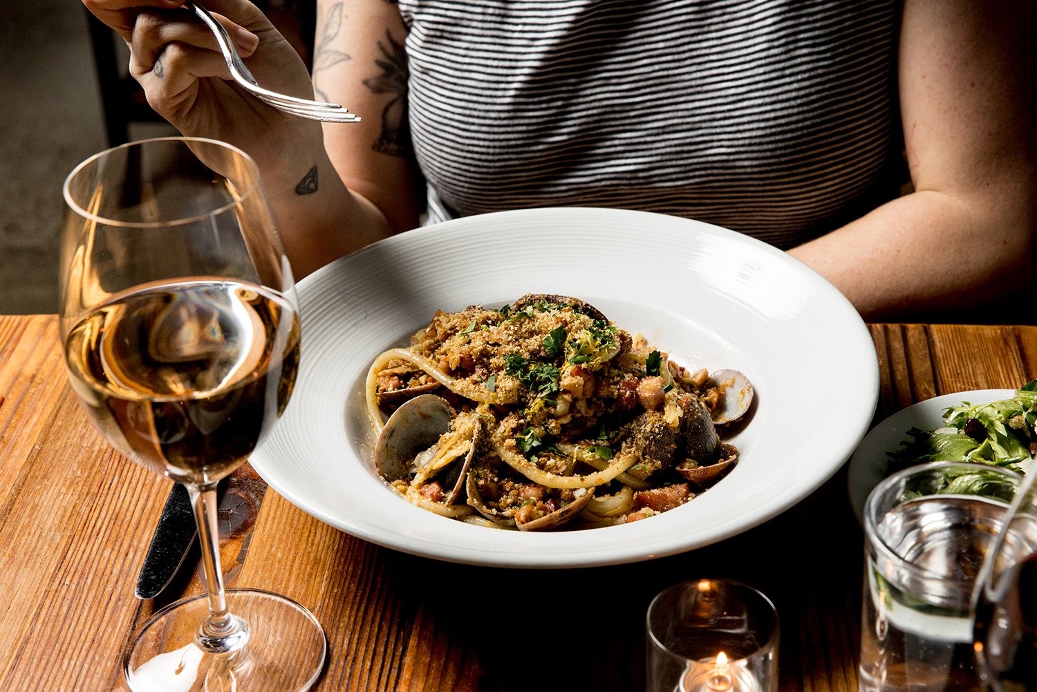 A person in a striped shirt is seated at a wooden table with a white plate of seafood pasta, a glass of white wine, and a side salad. The person's right arm and part of their torso are visible, with tattoos on their arm.
