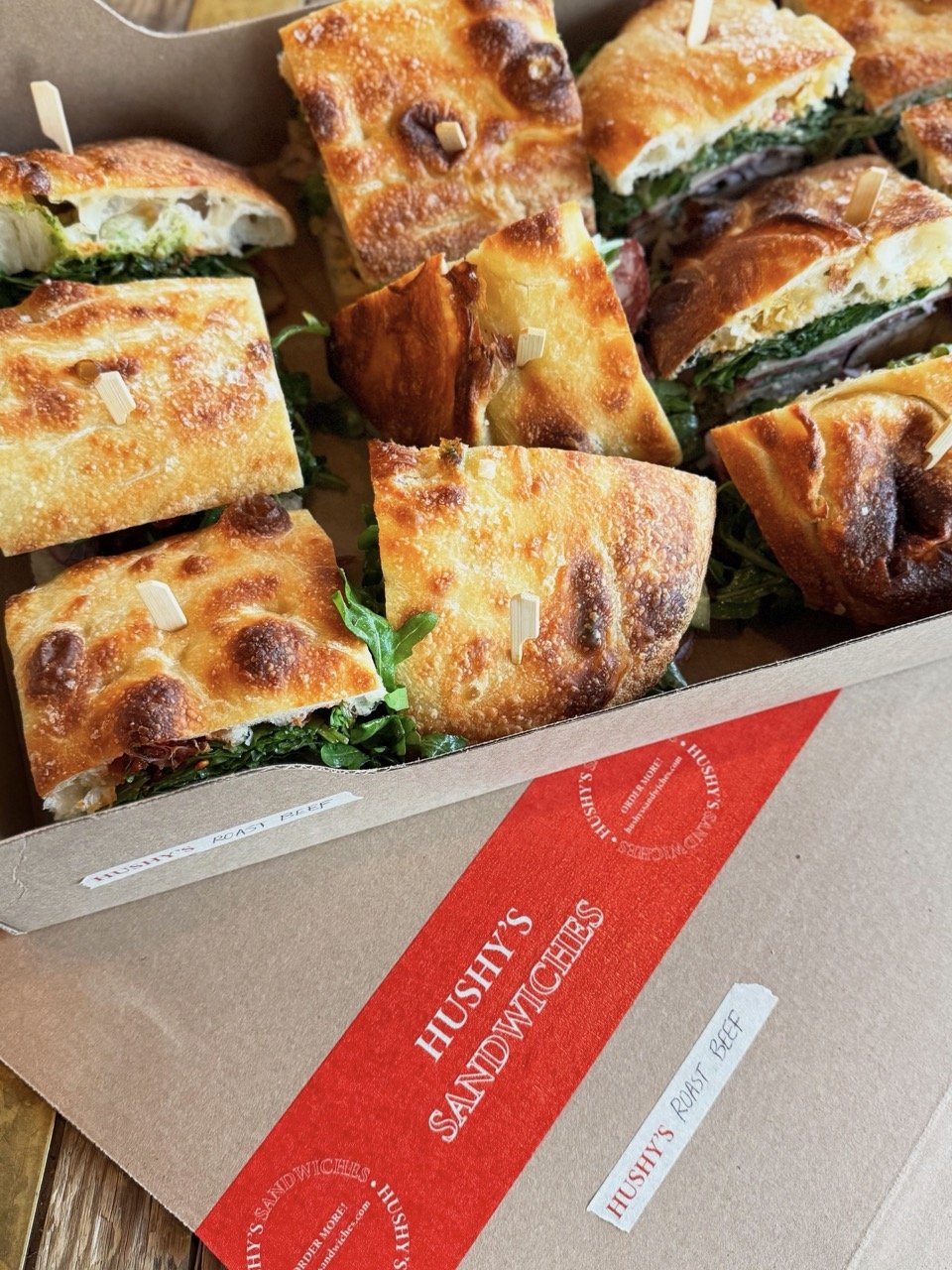 Close-up of a box of sandwiches from Hushy's Sandwiches, with breaded and baked sandwiches filled with roast beef, cheese, and greens, held together with small toothpicks.