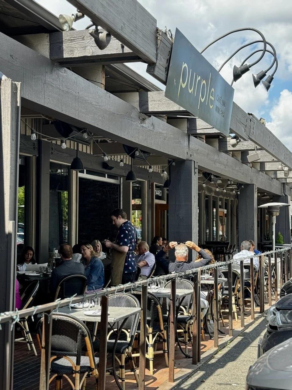 People dining outside a restaurant named Purple, with a sign and string lights, during daytime with a partly cloudy sky.