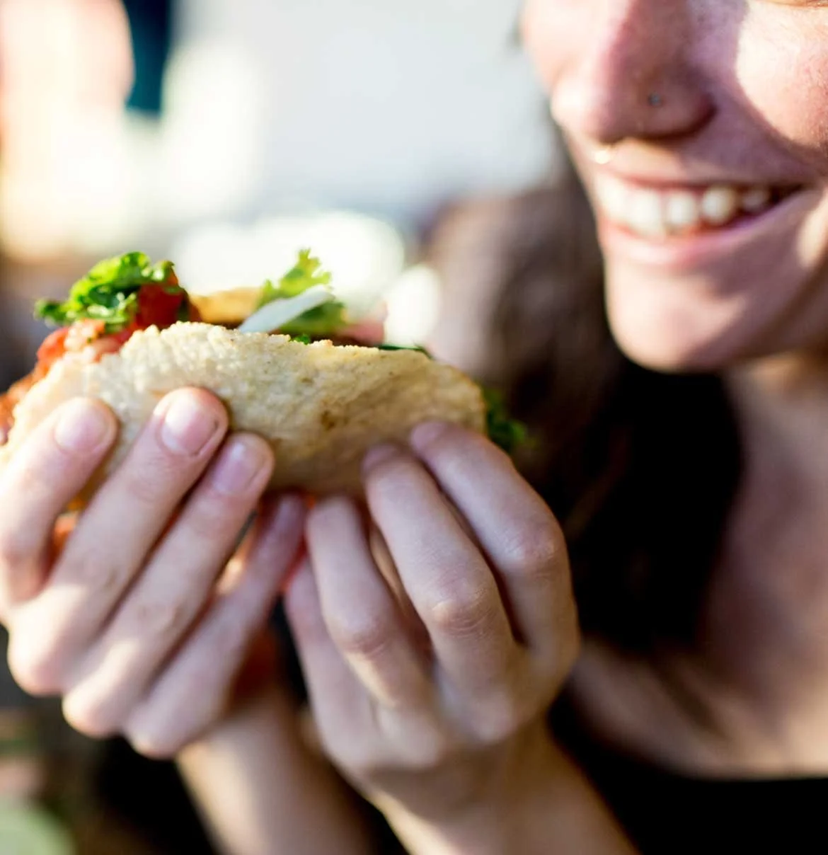 Person holding and about to bite into a sandwich with lettuce, tomato, and other fillings, smiling.