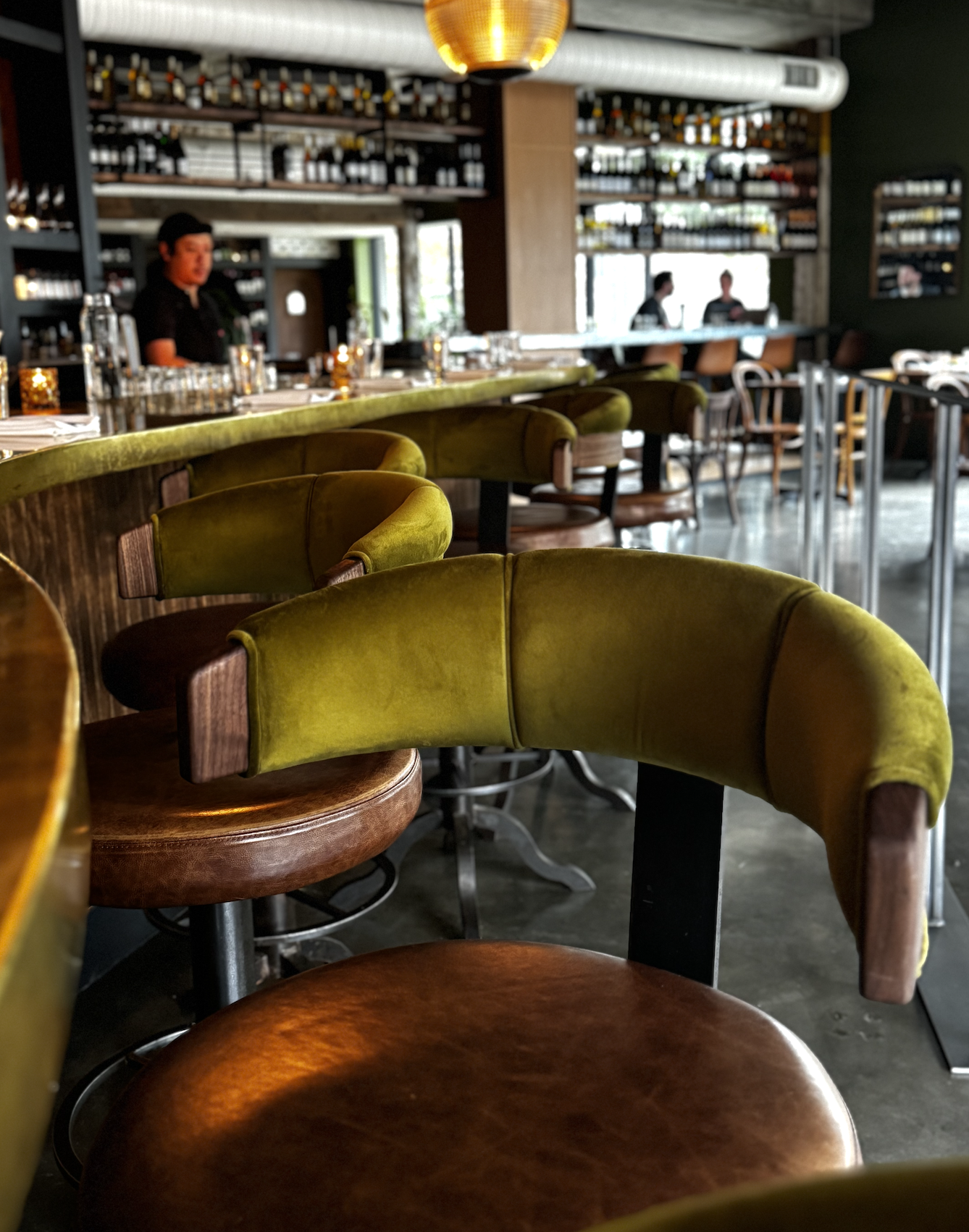 The image shows an empty restaurant with green and brown chairs, some with wooden finishes and some with velvet upholstery. In the background, there are shelves stocked with bottles, a bar area, and three staff members, two of whom are engaged in con