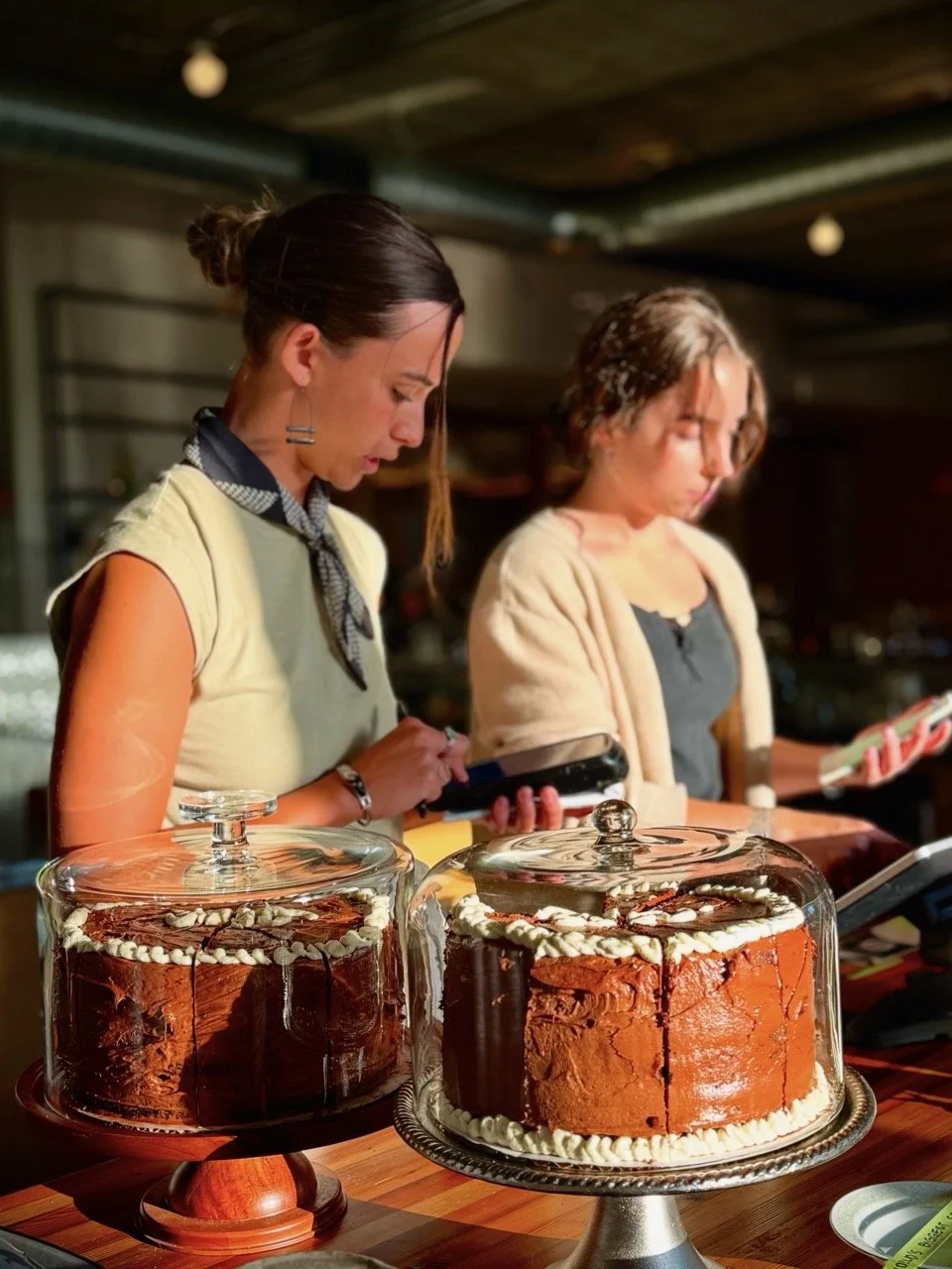 Two women working at a bakery or cafe, preparing desserts with two large cakes under glass covers on the counter in front of them.