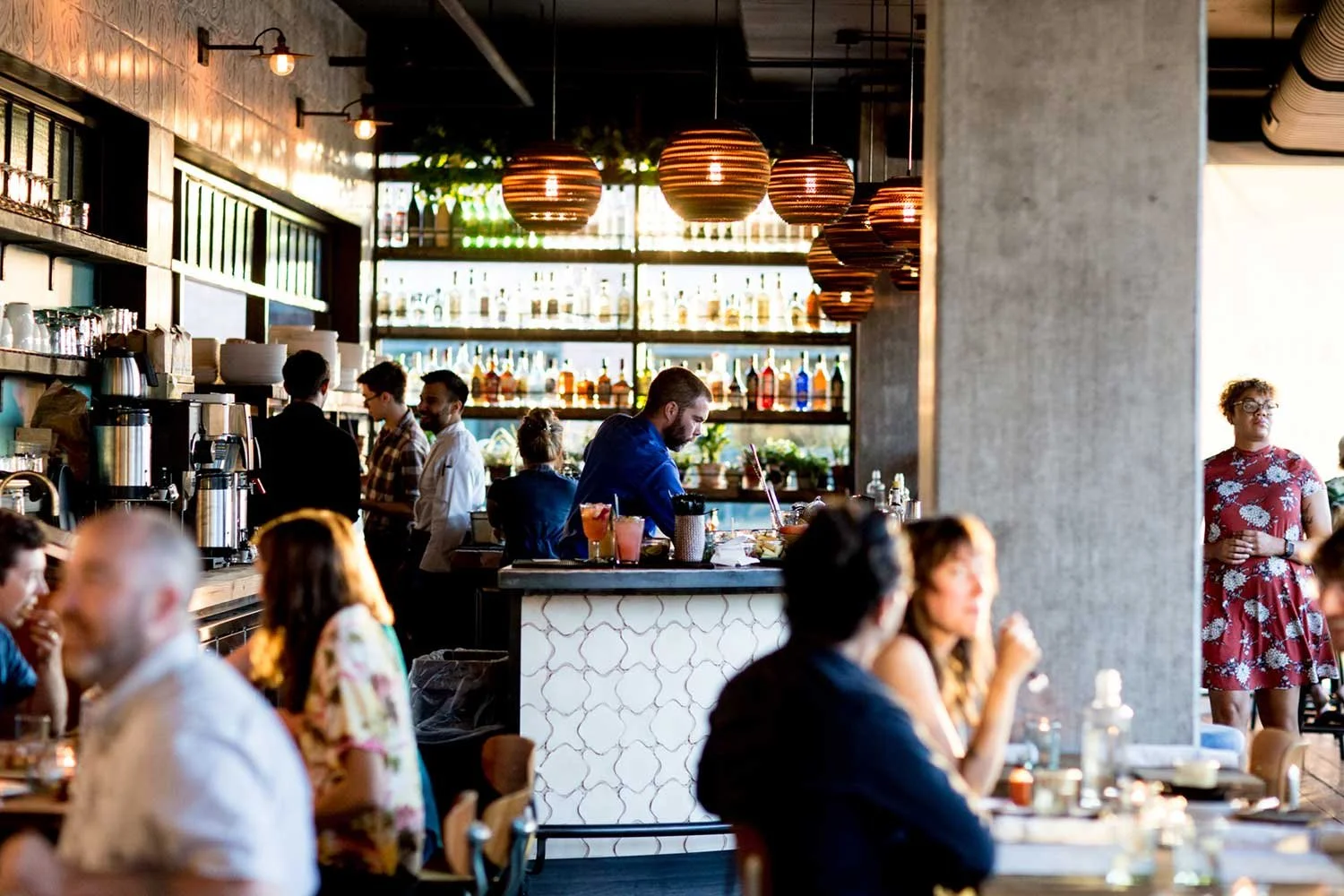 People dining and socializing in a modern restaurant with a bar, hanging pendant lights, and a busy atmosphere.