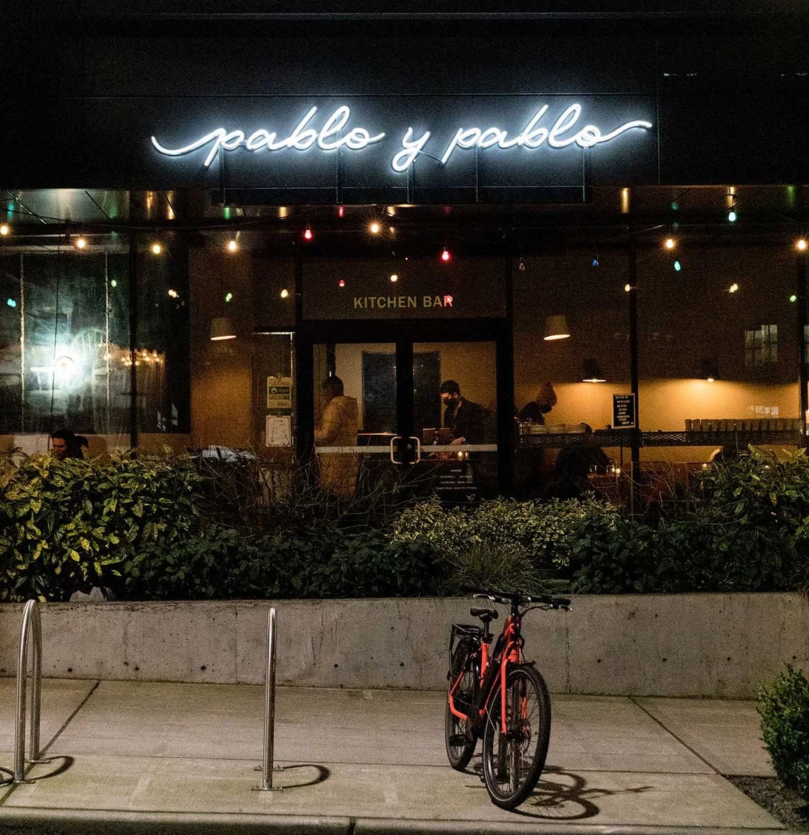 Night view of a restaurant named 'pablo y pablo' with a neon sign, interior visible through large windows showing people inside, a bike locked outside, and bushes in the foreground.