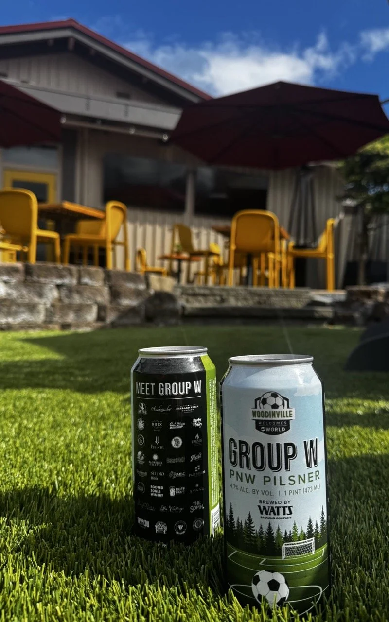 Two cans of beer sitting on green grass outside, with a patio area featuring yellow chairs and tables, and a building with a red roof and umbrellas in the background on a sunny day.