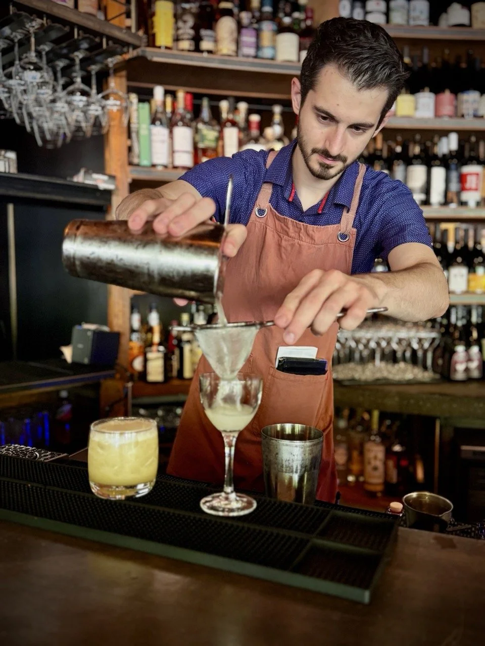 A bartender wearing a blue shirt and an apron pouring a cocktail into a glass at a bar, with various bottles of alcohol on shelves behind him.