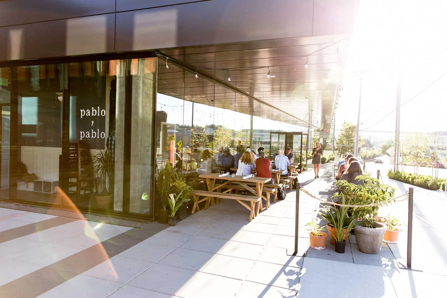 Outdoor seating area of a restaurant with people dining and a waitress serving, with potted plants and sunlight