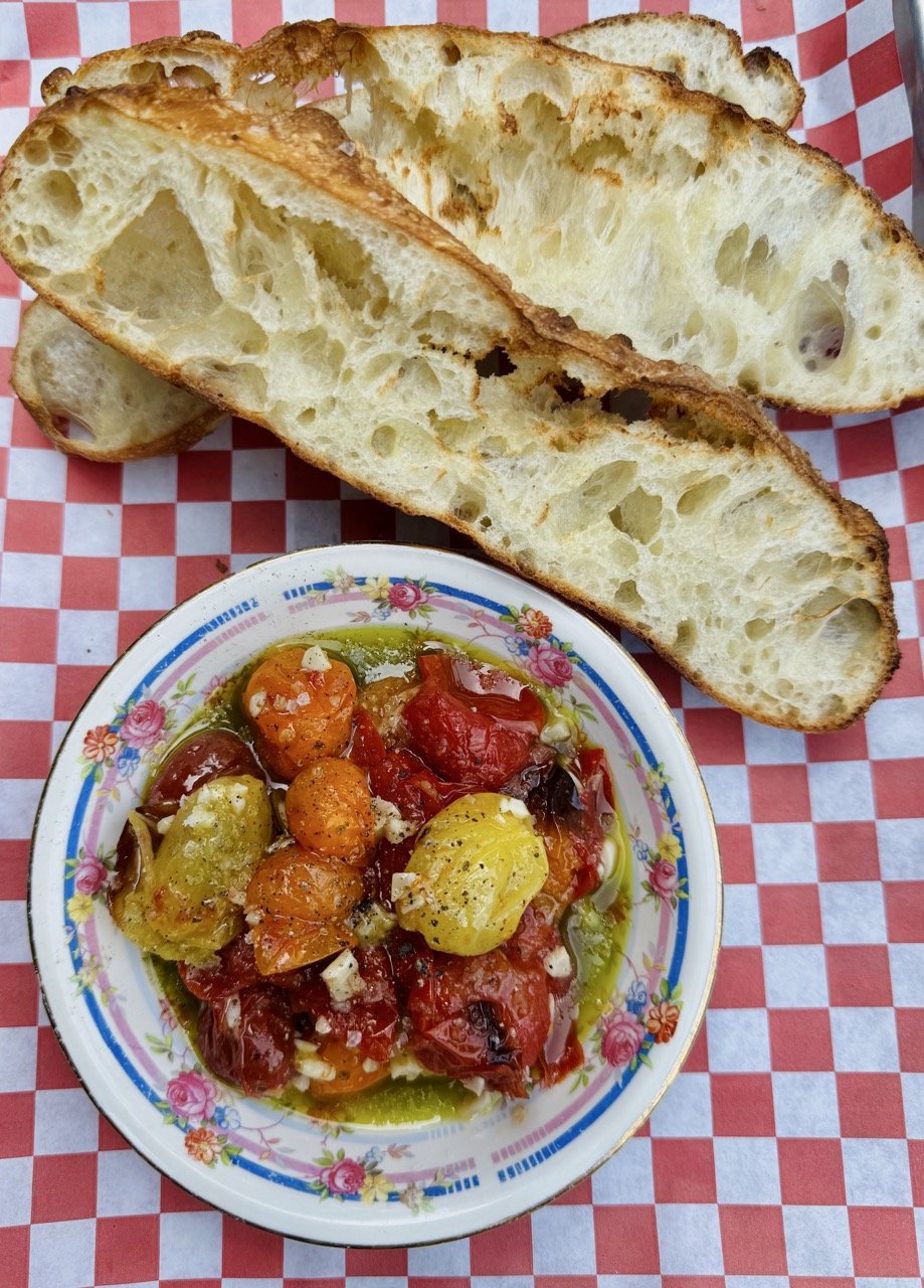 A bowl of roasted vegetables including tomatoes, carrots, and peppers, with bread slices placed on a red and white checkered tablecloth.