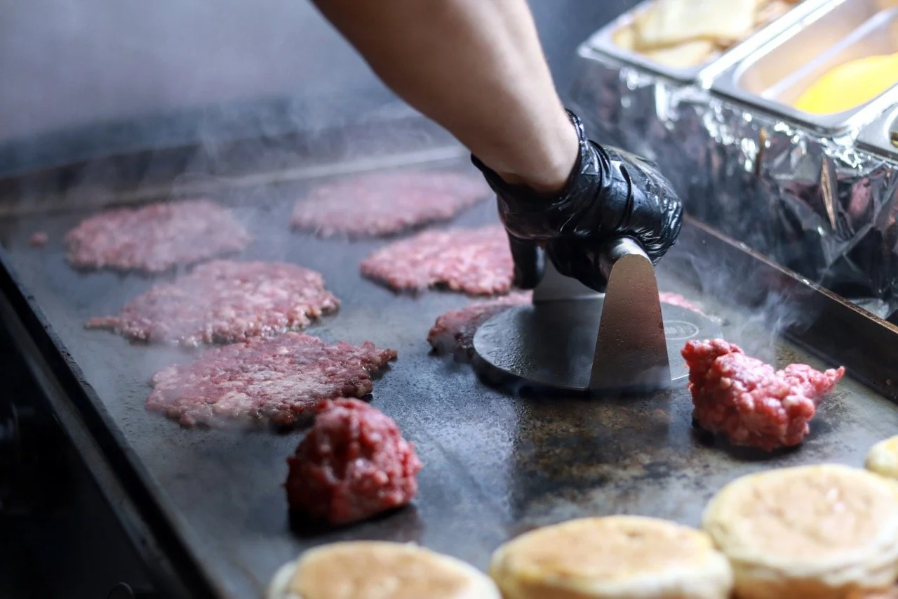 Person cooking hamburger patties on a flat grill, using a metal spatula, with various raw burger ingredients on the grill.