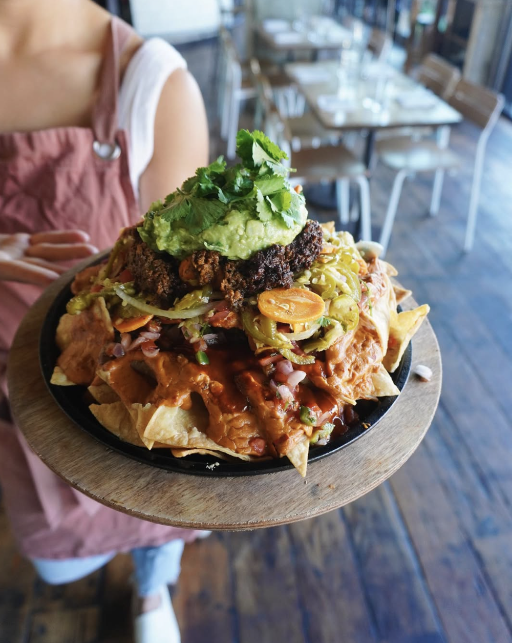 Person holding a plate of nachos topped with guacamole, sour cream, and cilantro in a restaurant.